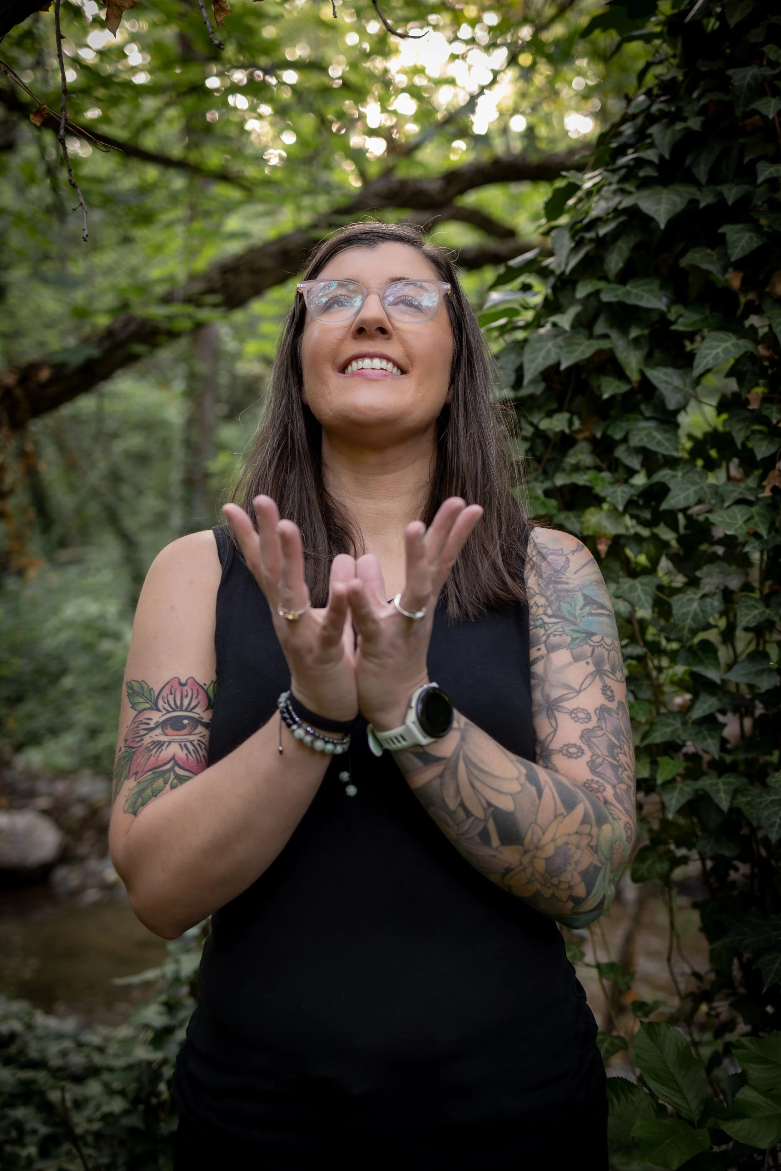 A woman with long dark hair and glasses, wearing a black sleeveless top, standing outdoors surrounded by green foliage, smiling and touching her hands together.