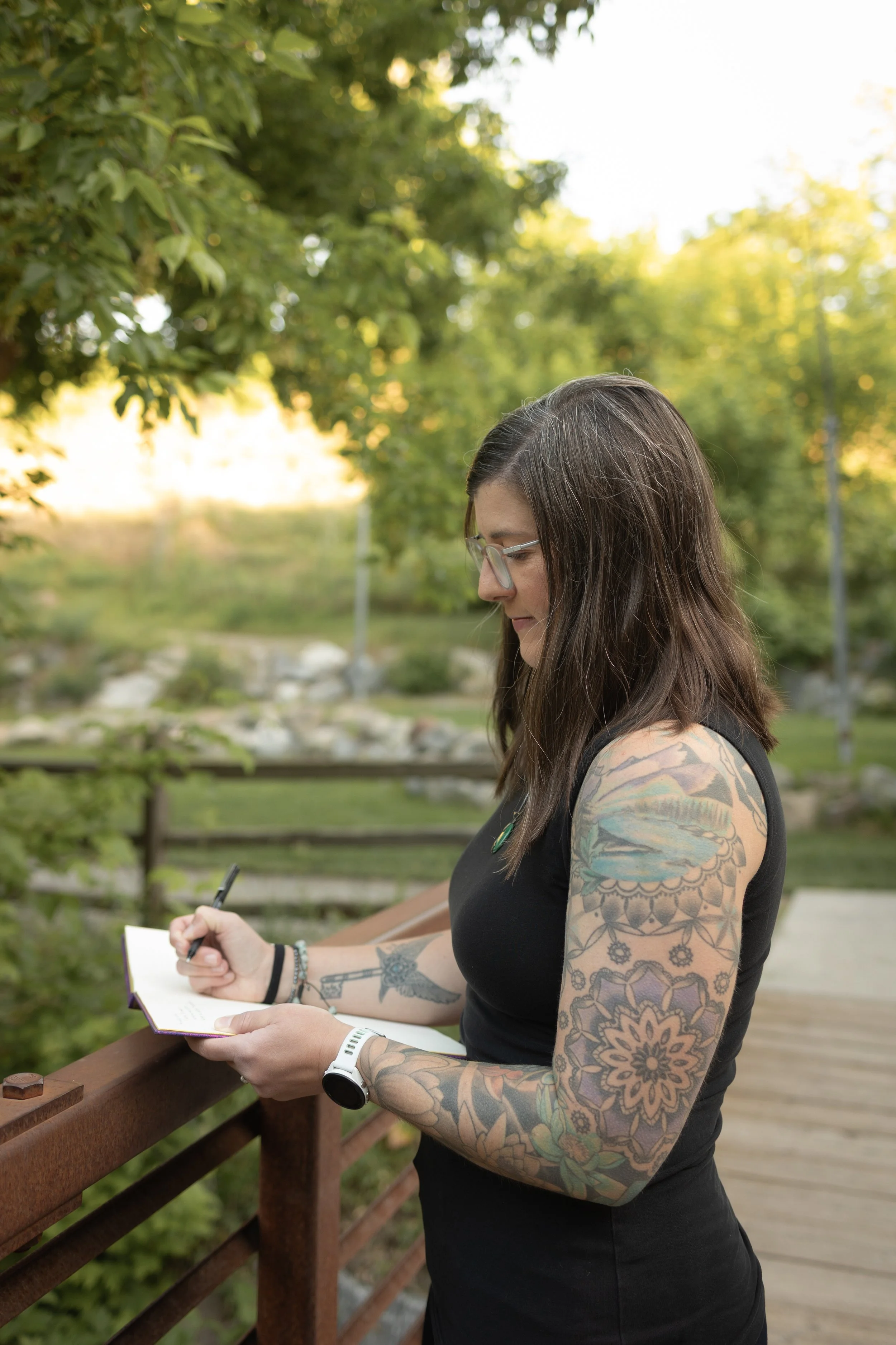A woman with long dark hair, glasses, and tattoos on her arms, standing on a wooden bridge in a lush, green park during sunset, writing in a notebook.