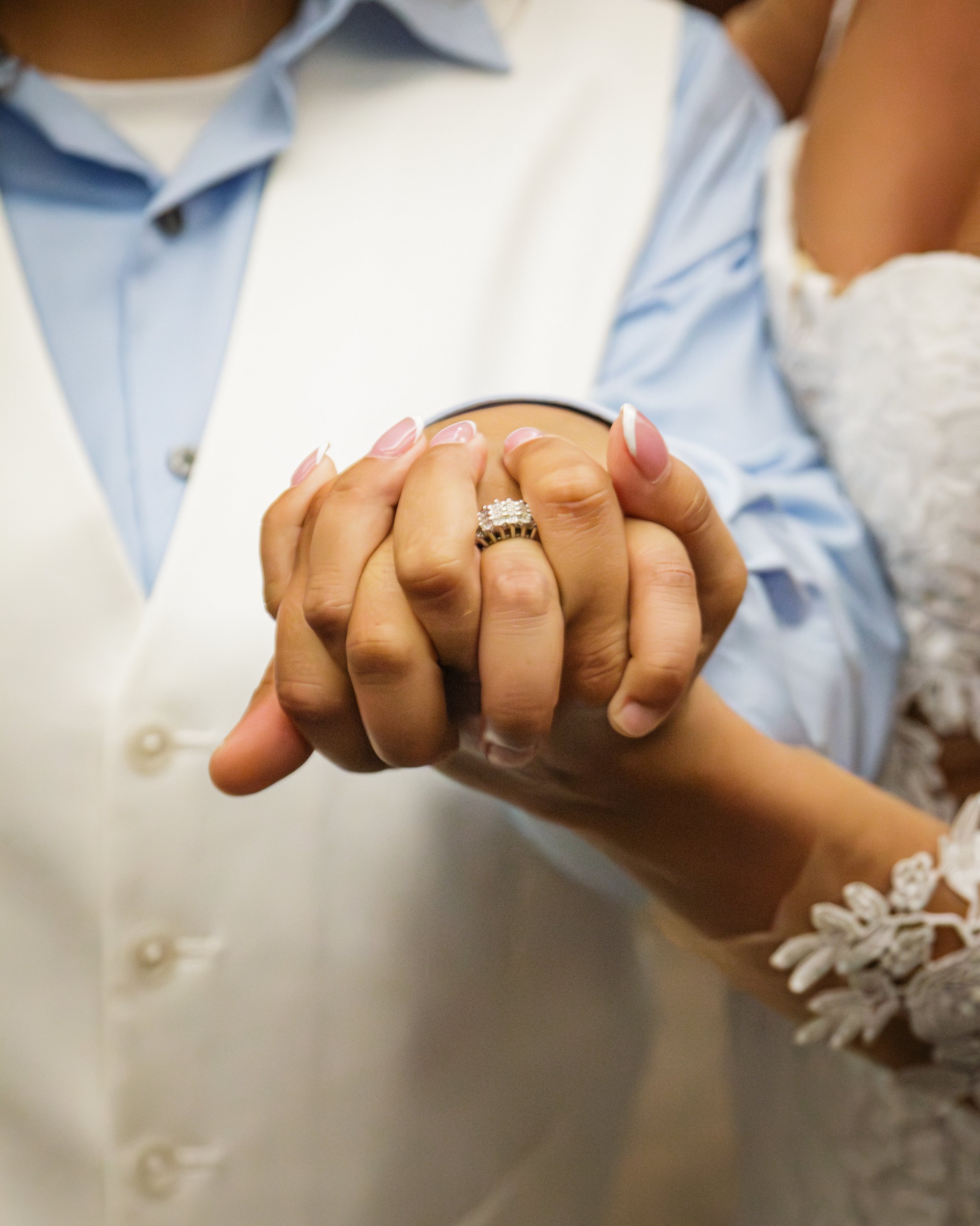 Close-up of a couple holding hands, displaying wedding rings, with the woman wearing a lace dress and a floral bracelet.