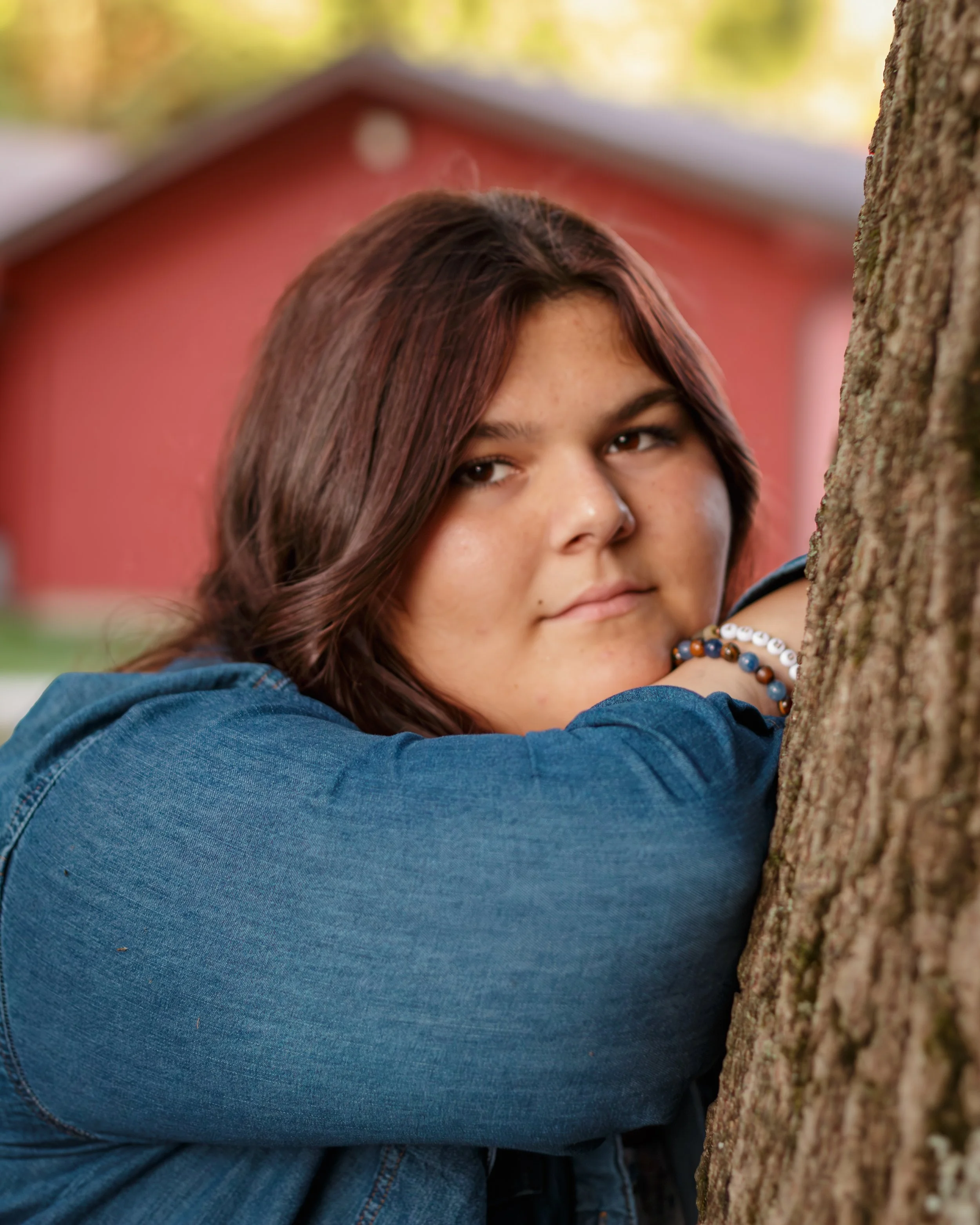 A woman with brown hair and a denim jacket leaning on a tree trunk outdoors.