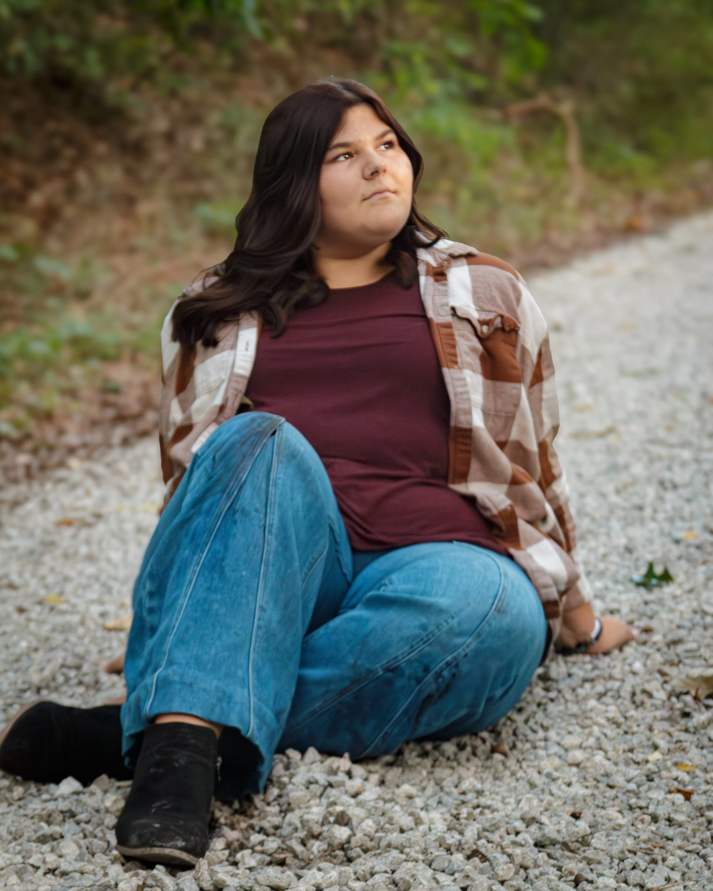 A young woman with dark hair sitting on a gravel path in a forest, wearing a camouflage jacket, maroon shirt, blue jeans, and black boots.