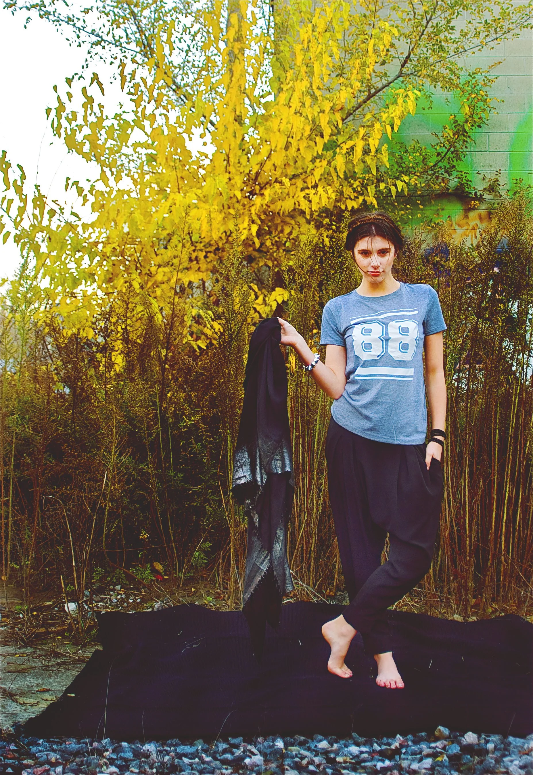 A young woman standing barefoot on a black cloth outdoors, holding a black scarf in her left hand, surrounded by yellow foliage and tall grasses.