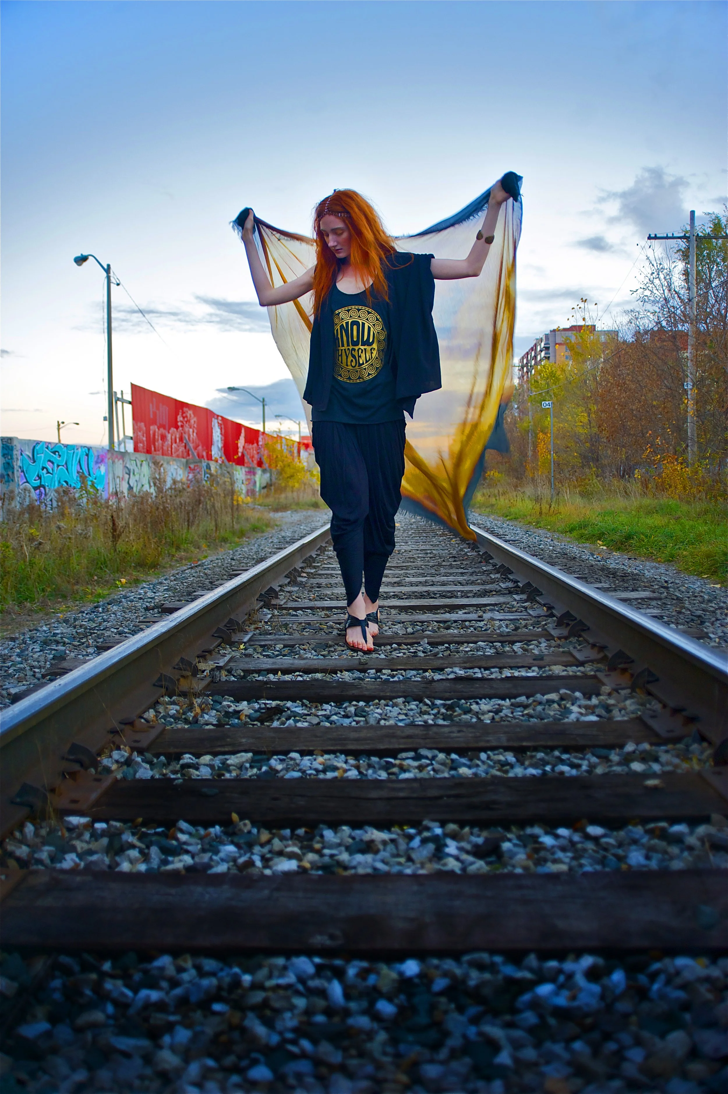 A woman with long red hair walking barefoot on railroad tracks at sunset, holding a sheer cloth behind her with graffiti-covered walls on one side and trees on the other.