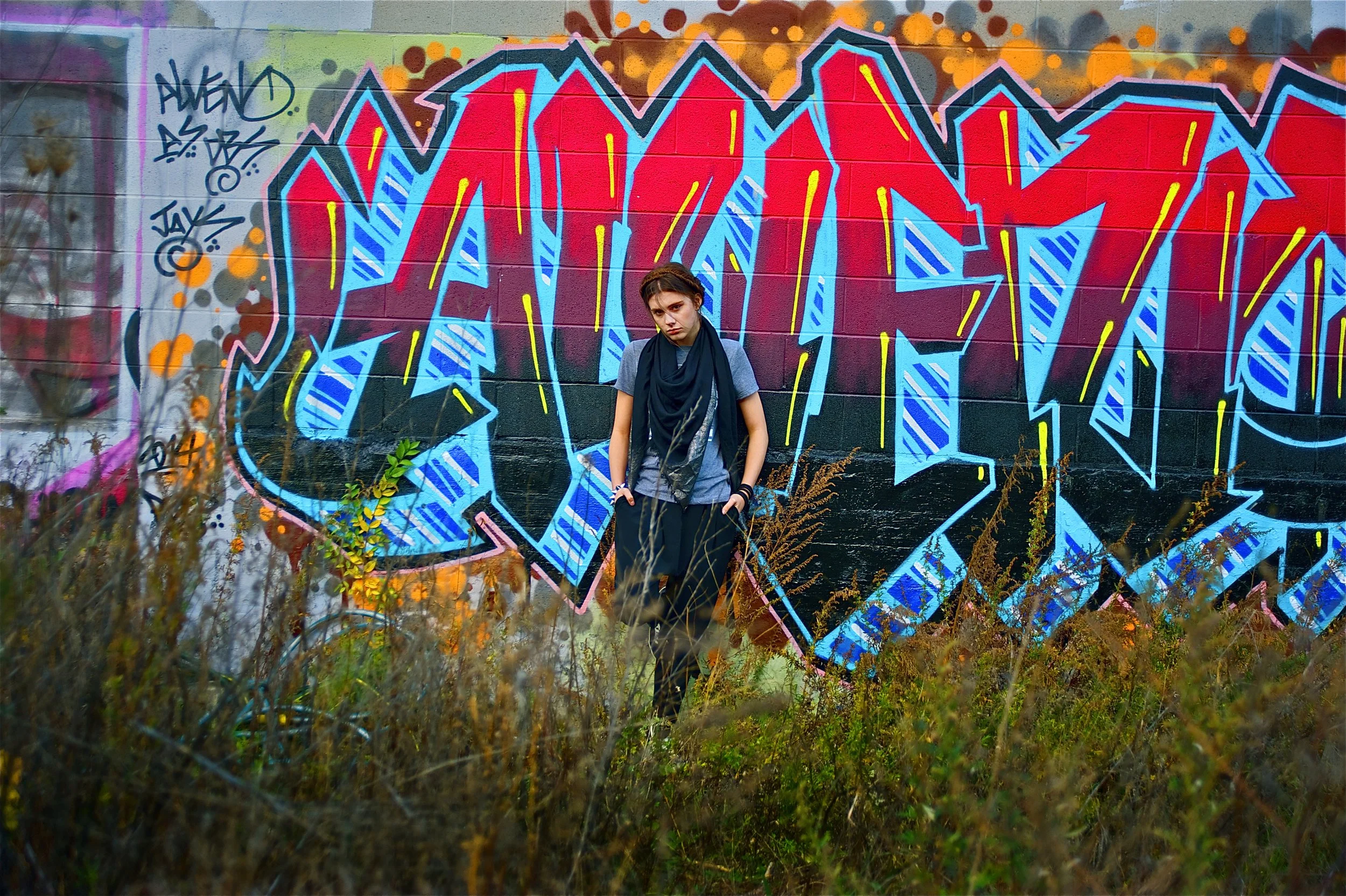 Young woman with short hair standing in front of colorful graffiti art on a brick wall, surrounded by overgrown grass and weeds.