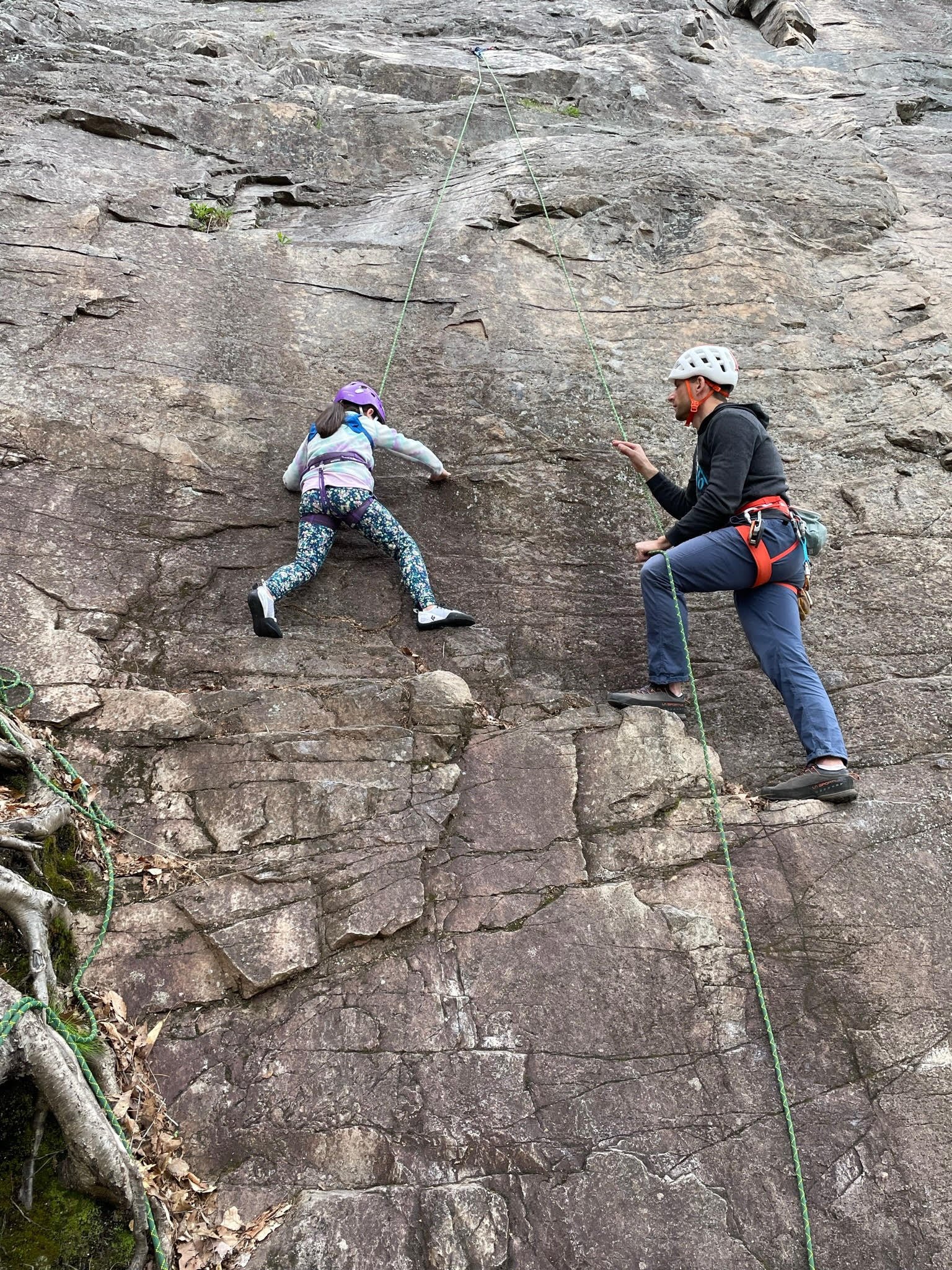A child and an adult rock climbing on a cliff, both wearing helmets and harnesses. The child is climbing while the adult is belaying, holding the rope for safety.