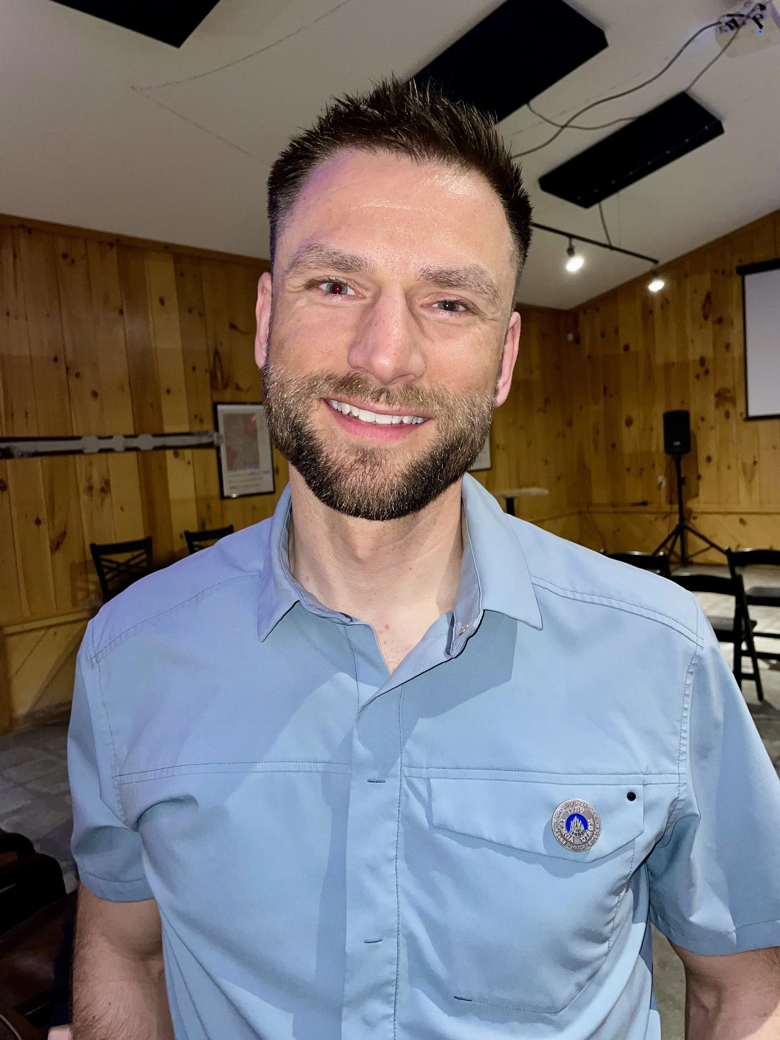 A man with a beard smiling, wearing a light blue shirt, standing indoors with wooden walls and ceiling lights visible in the background.