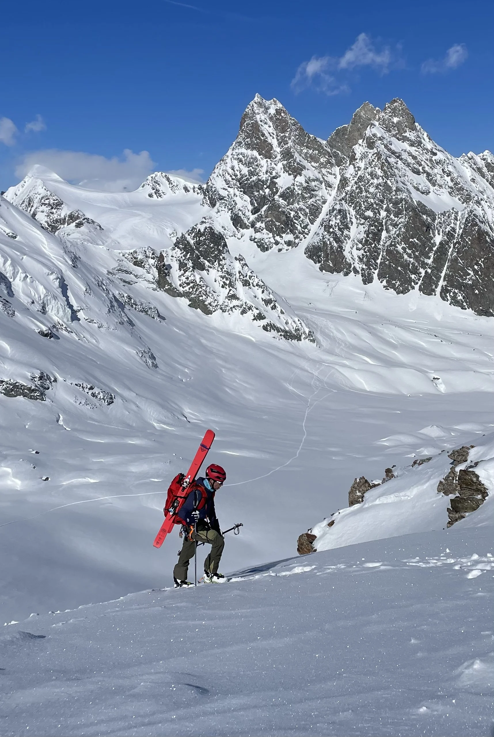 A person wearing a red helmet and carrying skis on their back is climbing a snowy mountain under a clear blue sky.