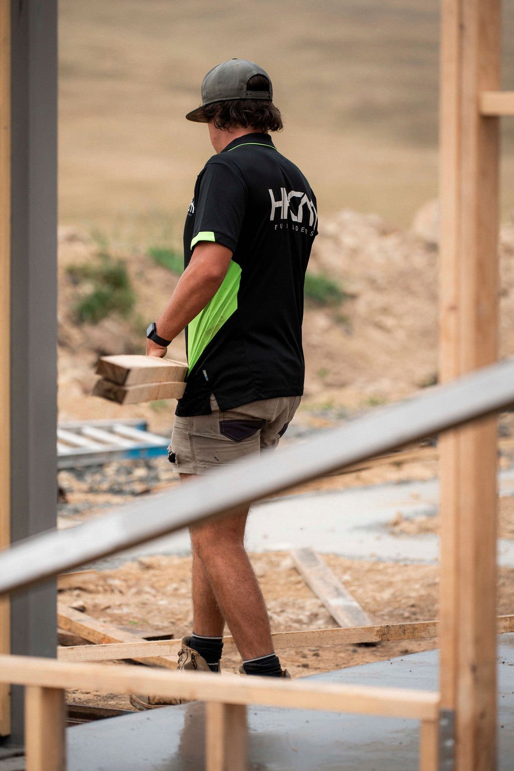 A man wearing a black and green shirt, shorts, and a cap is holding wooden boards at a construction site outdoors.