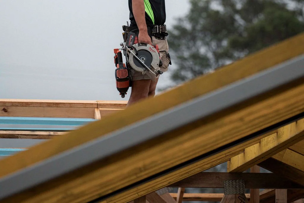 A construction worker standing on a building frame holding a power saw, wearing work shorts and a safety tool belt.