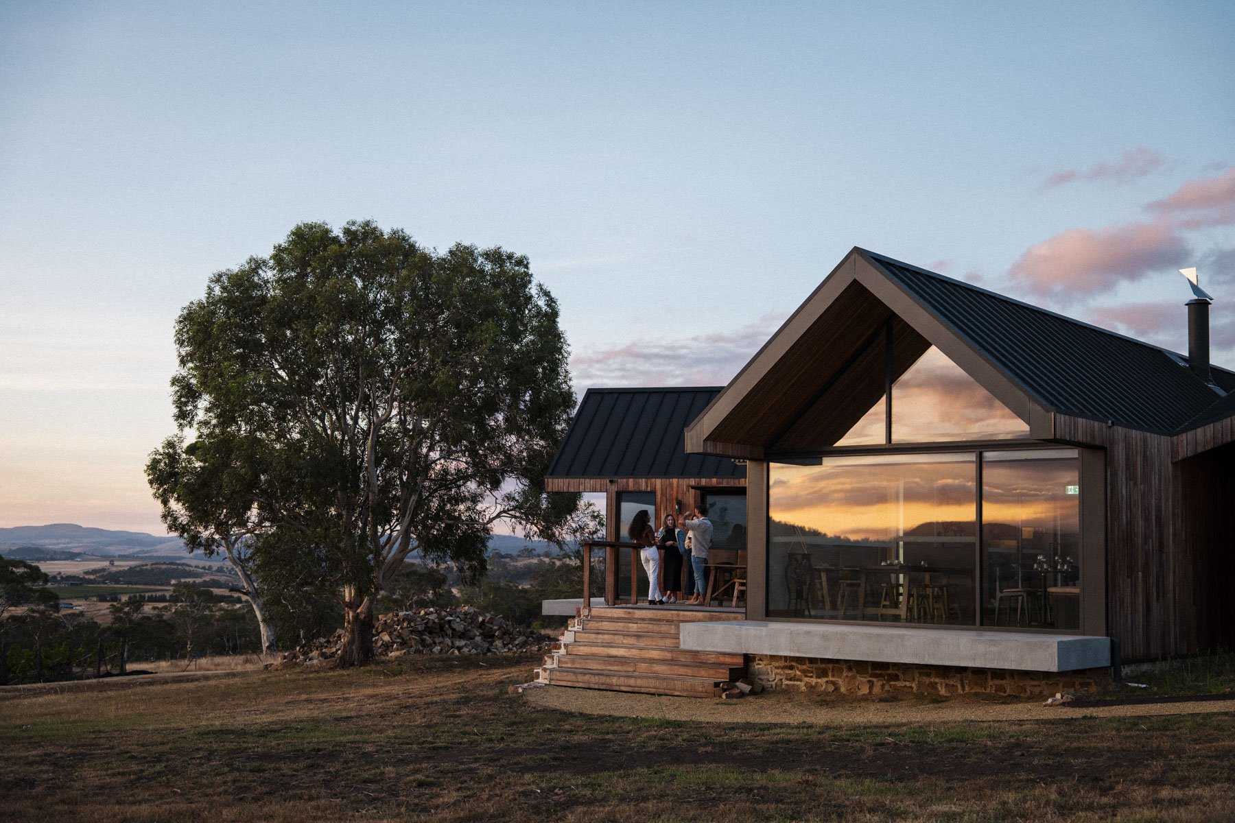 A modern house with a wooden exterior and large glass windows reflecting a sunset sky, situated on a grassy landscape with a large tree nearby.