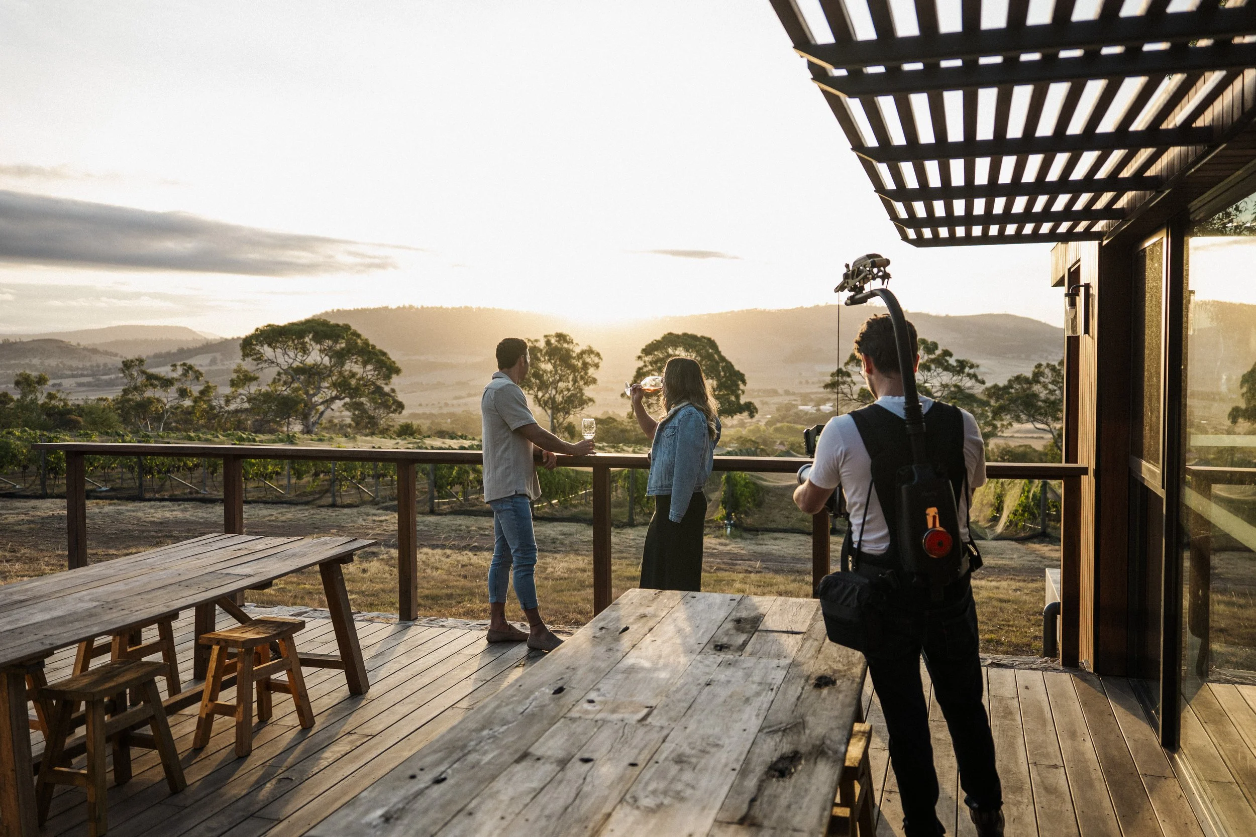 Three people on a deck with a sunset view overlooking trees and hills, two women sipping wine and a man nearby, with a camera operator filming them.