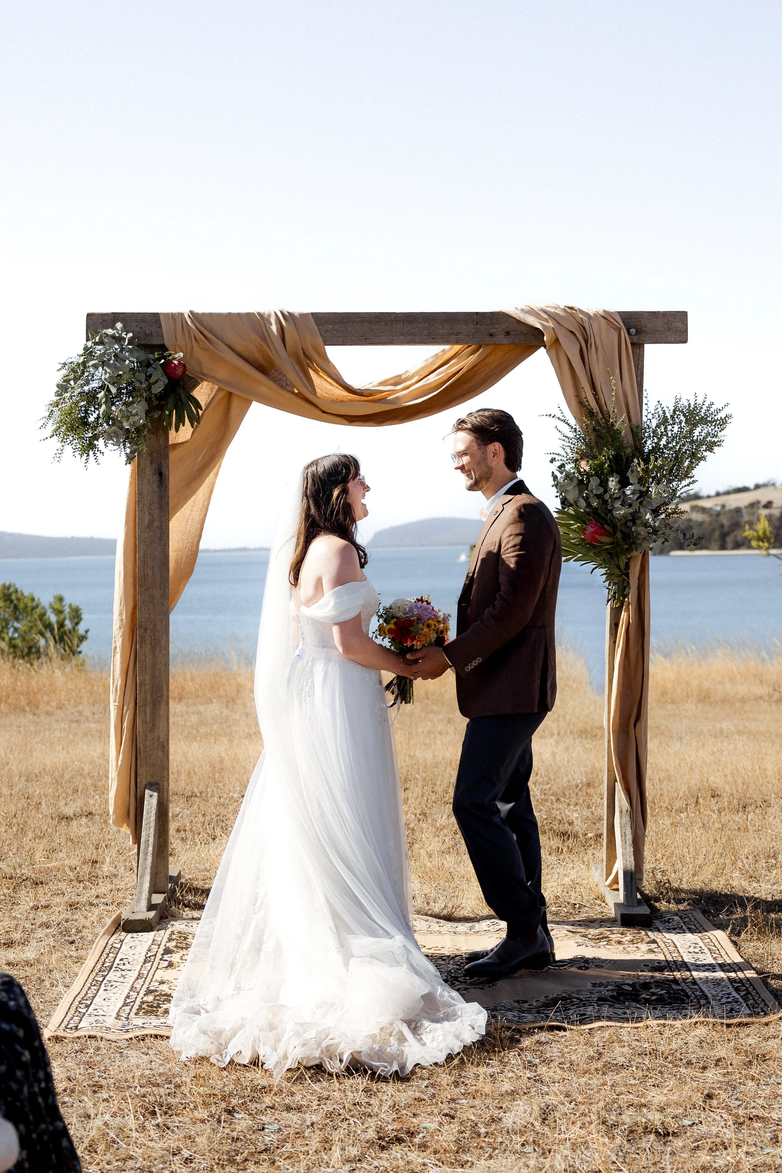 A bride and groom stand facing each other under an outdoor wedding arch decorated with beige fabric and greenery near a body of water. The bride is wearing a white off-shoulder wedding dress, and the groom is in a brown suit. They are smiling at each other, holding hands, and holding a bouquet of flowers. The scene is set on a grassy area with a calm lake and hills in the background, under a clear sky.