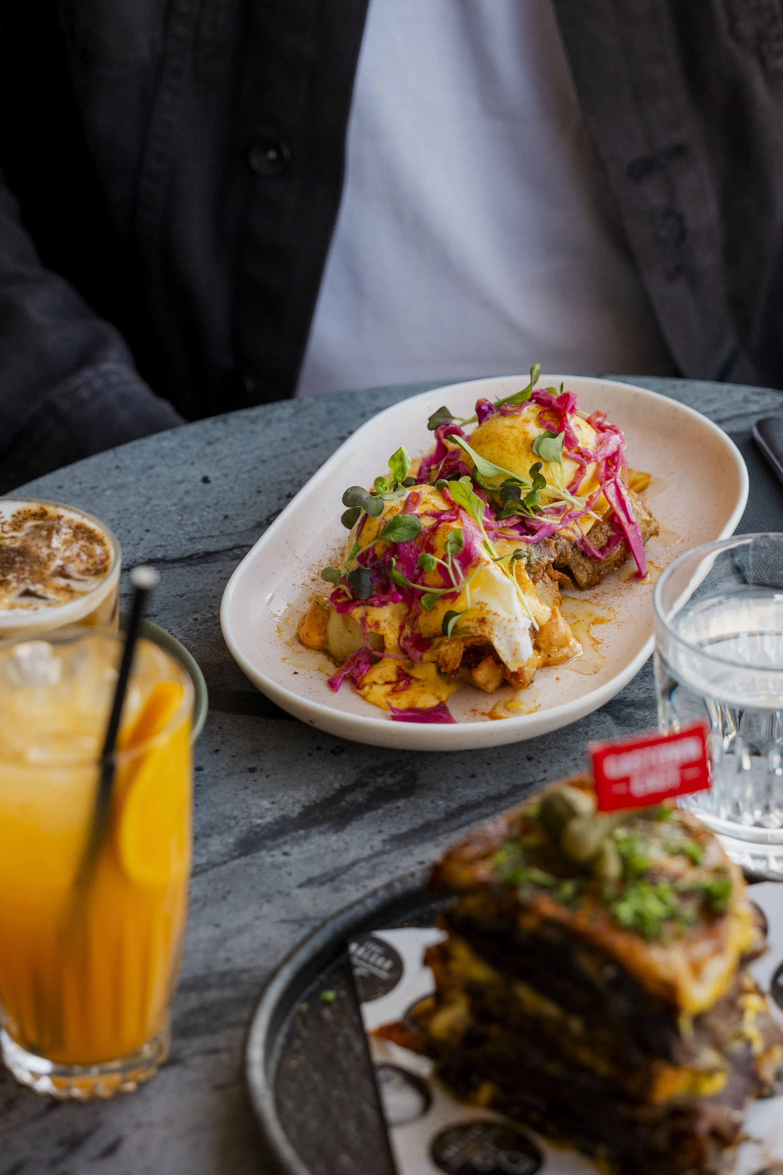 A white oval plate with eggs Benedict, topped with hollandaise sauce, herbs, and pink cabbage. Other dishes, drinks, and a glass of water are partially visible on the table.