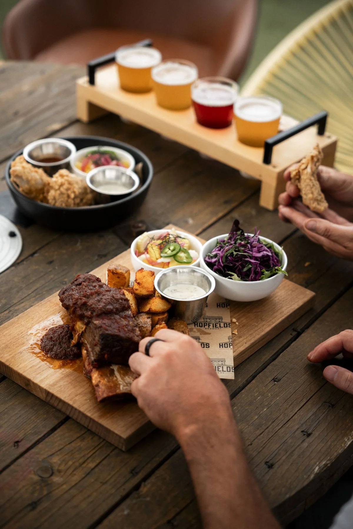 A rustic wooden table with a meal including a large cut of barbecued ribs, roasted potatoes, colorful salads, and dipping sauces on a wooden serving board. In the background, there are additional side dishes and drinks.