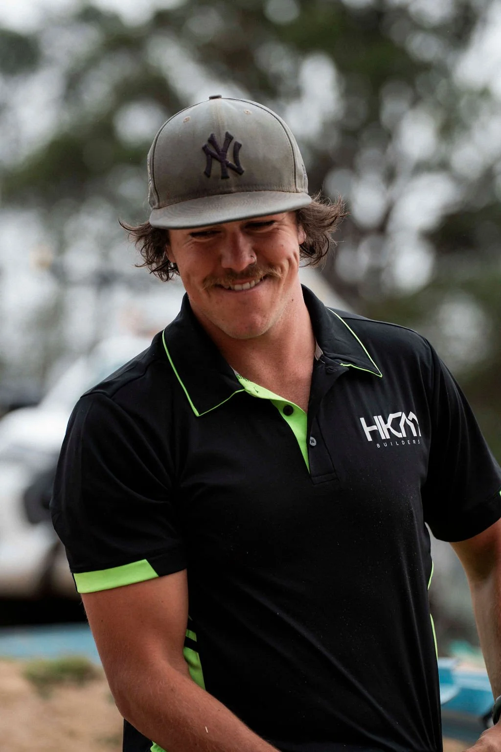 A smiling man with a mustache and wearing a New York Yankees cap and a black shirt with green accents and 'HKM Builders' logo.