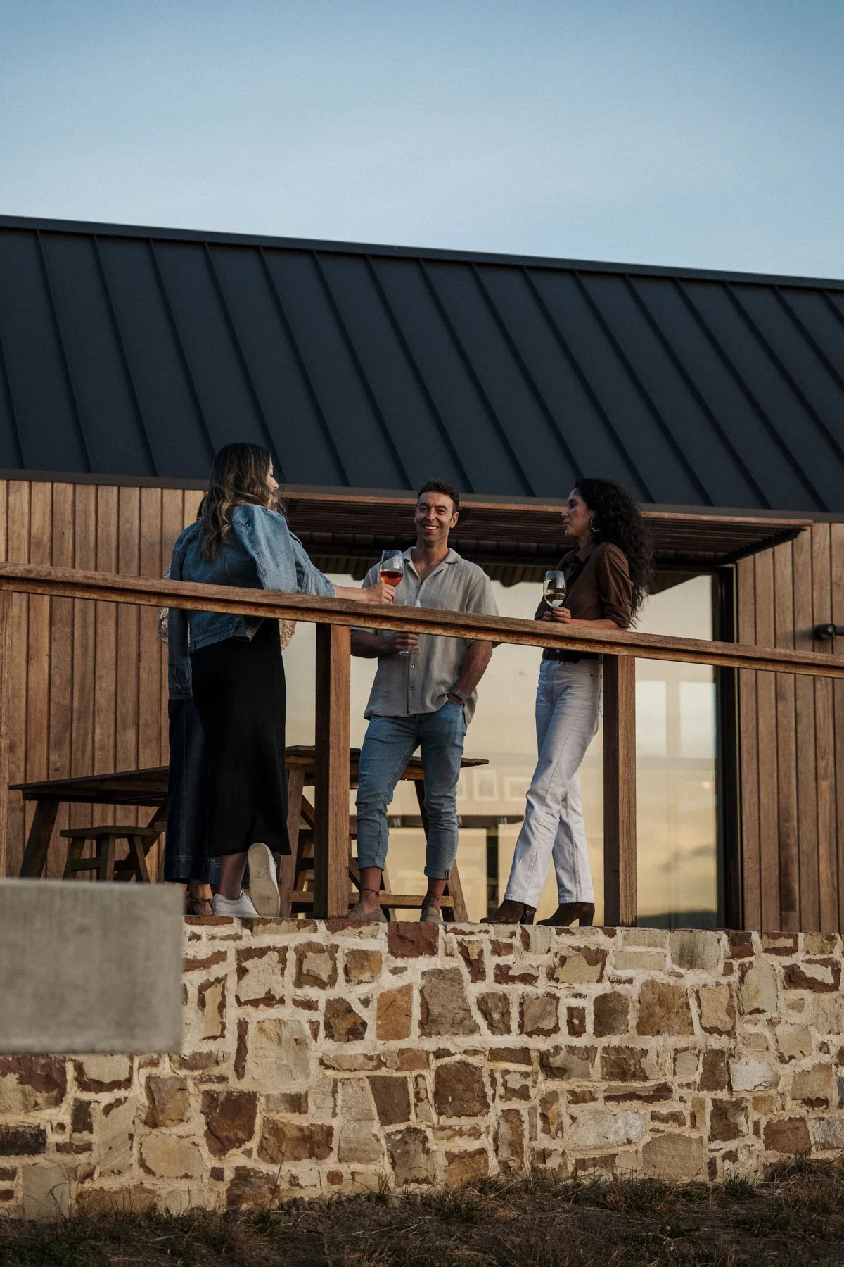 Three friends are standing on a wooden balcony, enjoying drinks and chatting on a warm evening.
