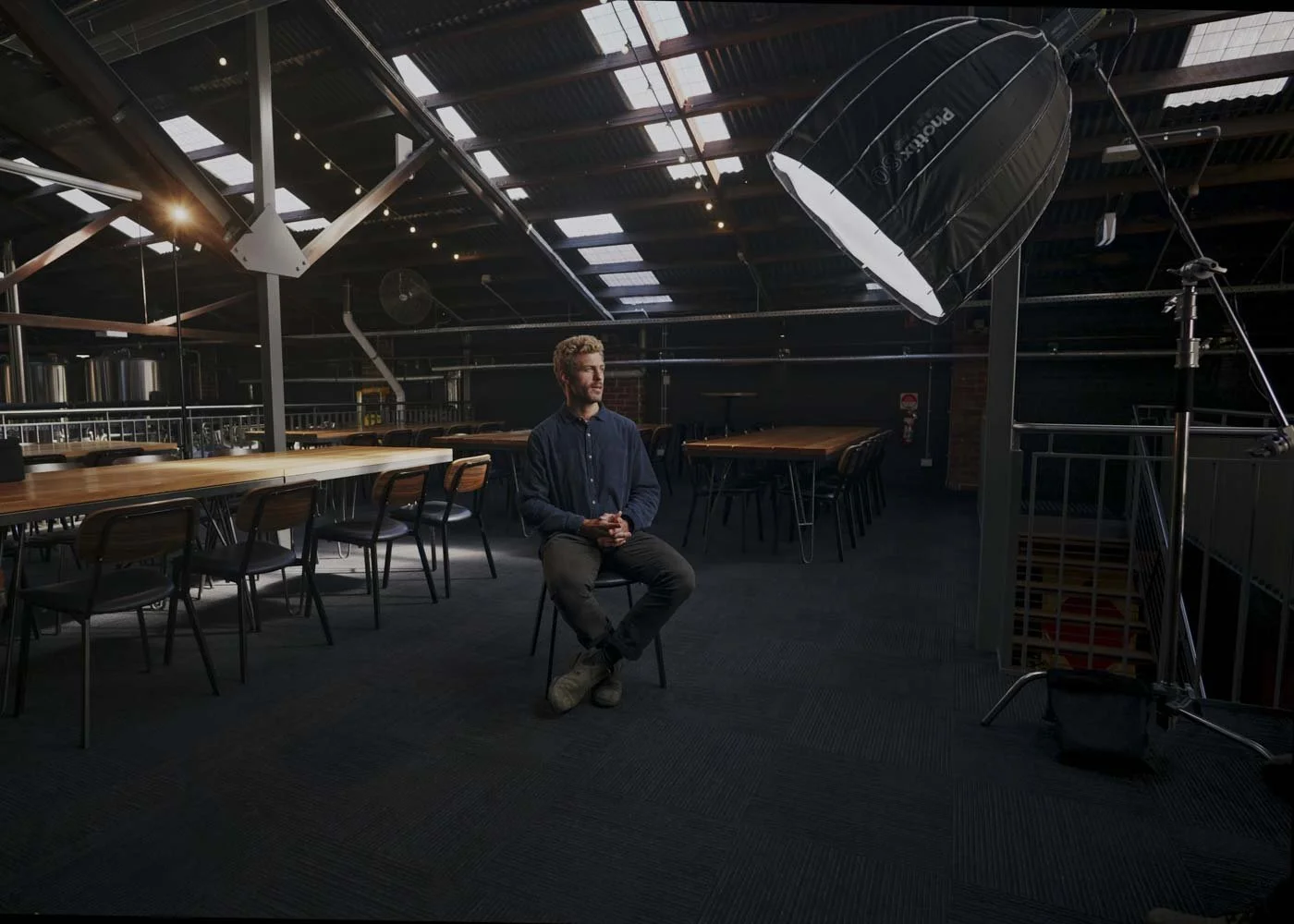 A man seated on a chair in an industrial-style space with large tables, hanging lights, and a professional photography lighting setup with a softbox.
