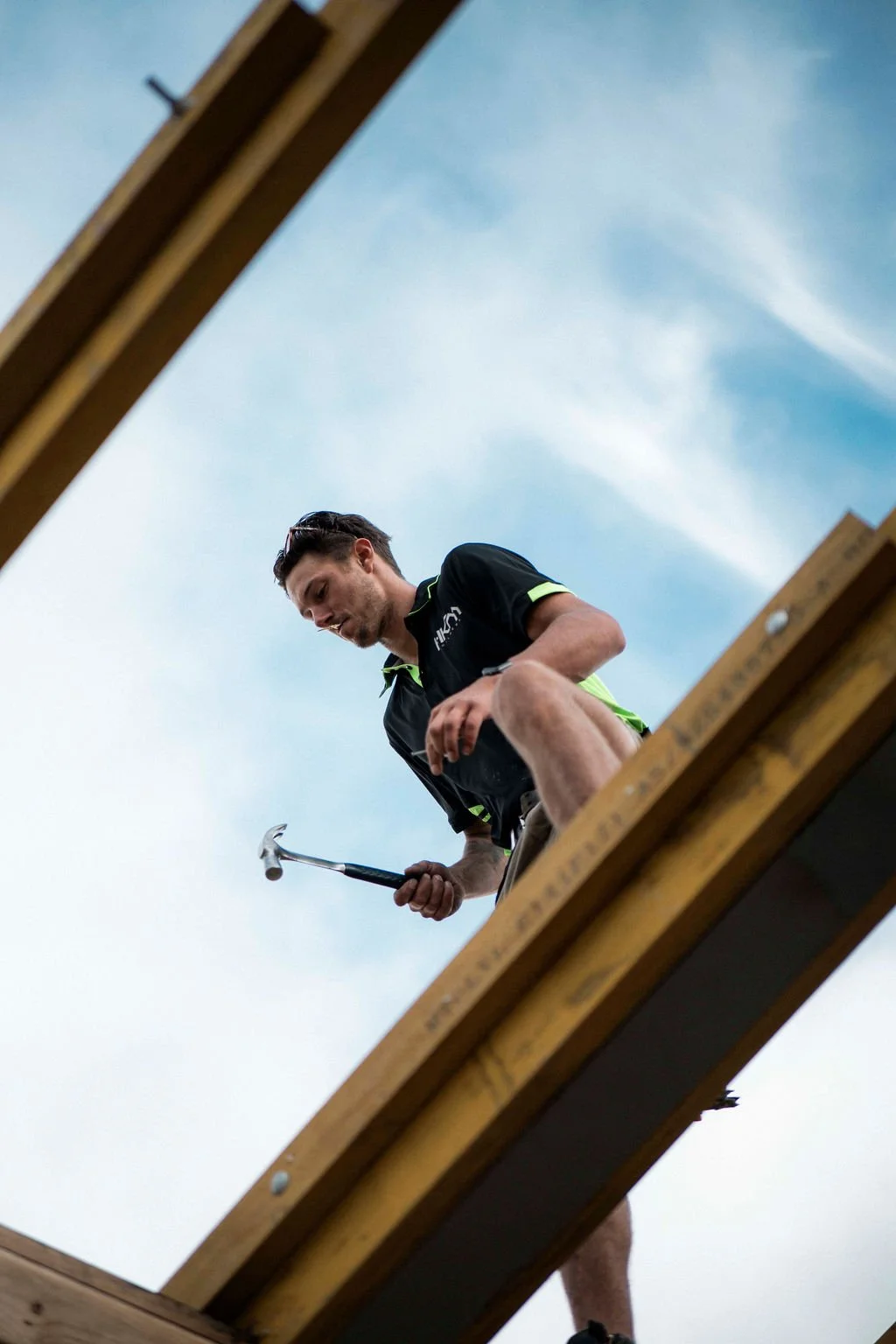 A man in athletic clothing peering down while holding a hammer, seen from below through an opening in a wooden structure against a blue sky.