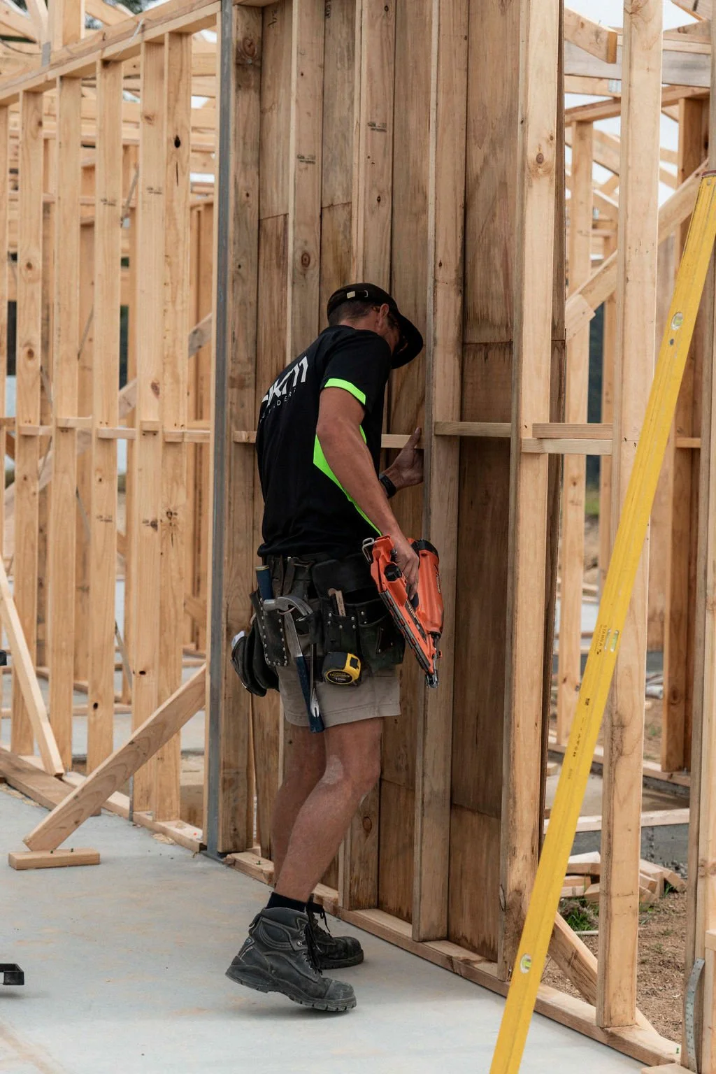 A construction worker inspecting or working on a wooden wall frame at a building site, holding a nail gun and wearing a tool belt.