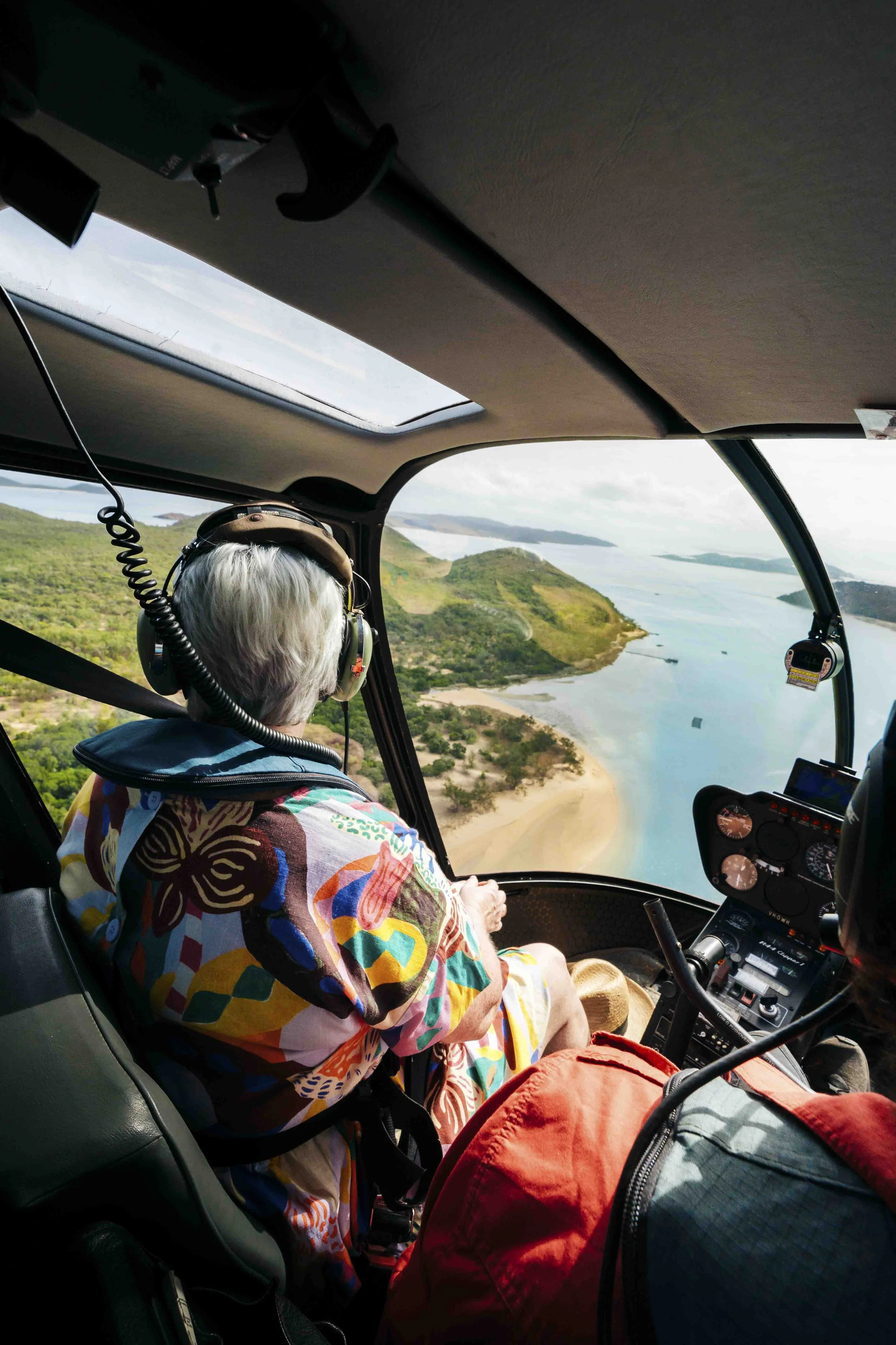 Aerial view from inside a helicopter cockpit showing an elderly person with white hair wearing headphones and a colorful shirt, flying over a scenic landscape with hills, water, and a beach.