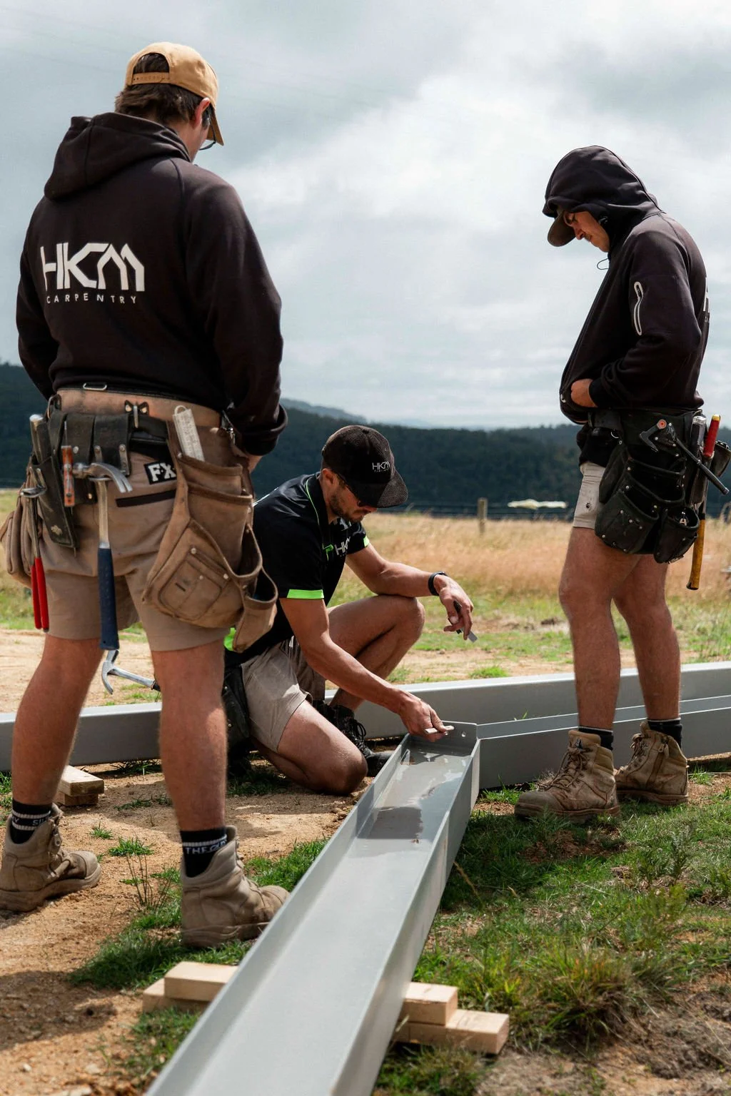 Three workers in outdoor gear installing a metal rainwater collection channel in a grassy field under cloudy skies.