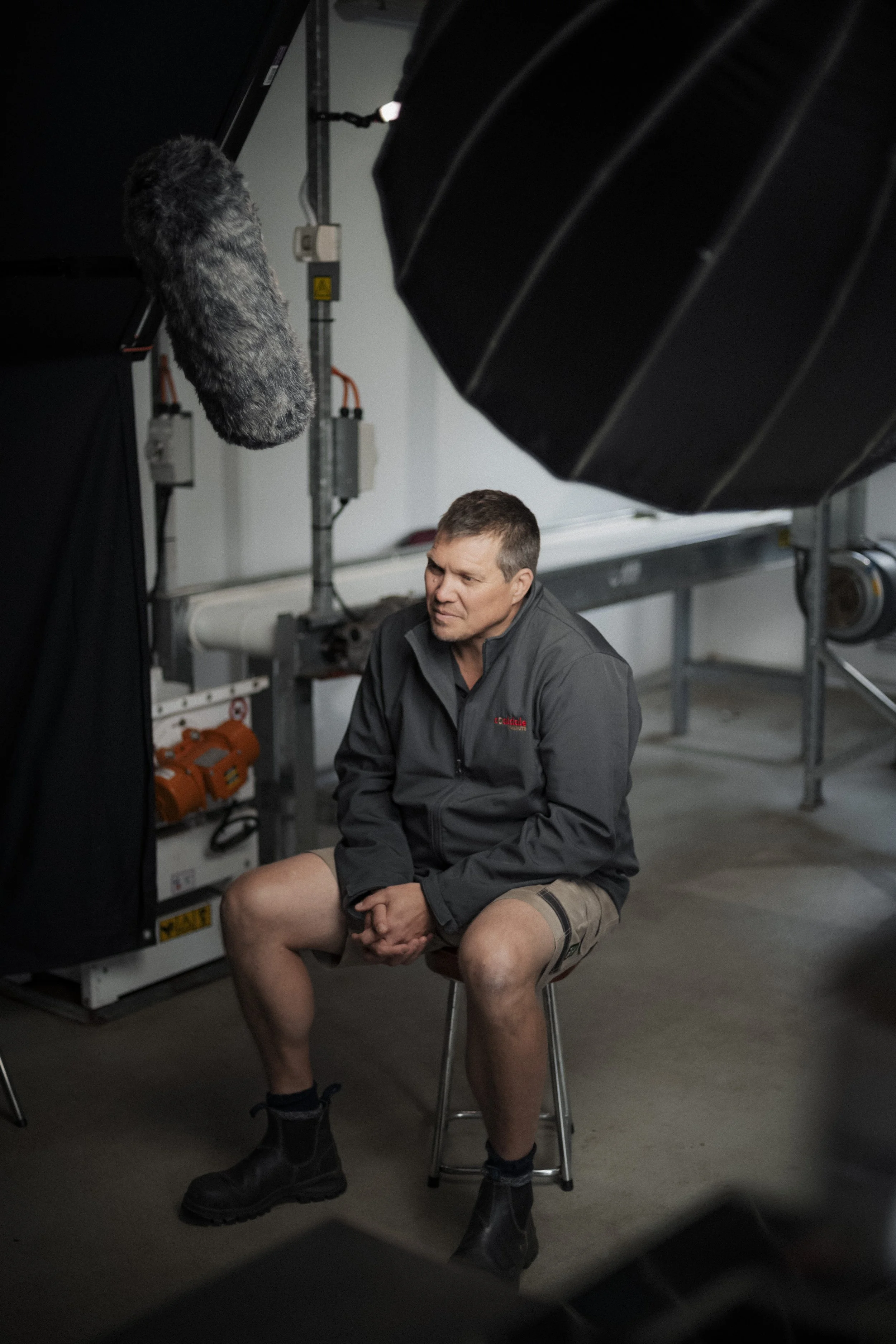 A man is sitting on a stool in a studio setting with professional lighting equipment and a microphone overhead, possibly being interviewed or filmed.