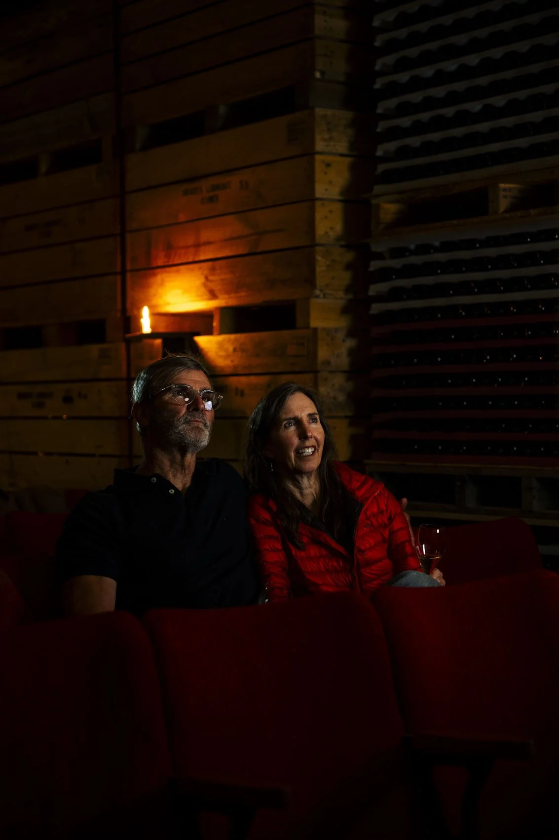 A man and a woman sitting together on a red couch in a dimly lit room with wooden walls, watching something with expressions of interest and surprise.