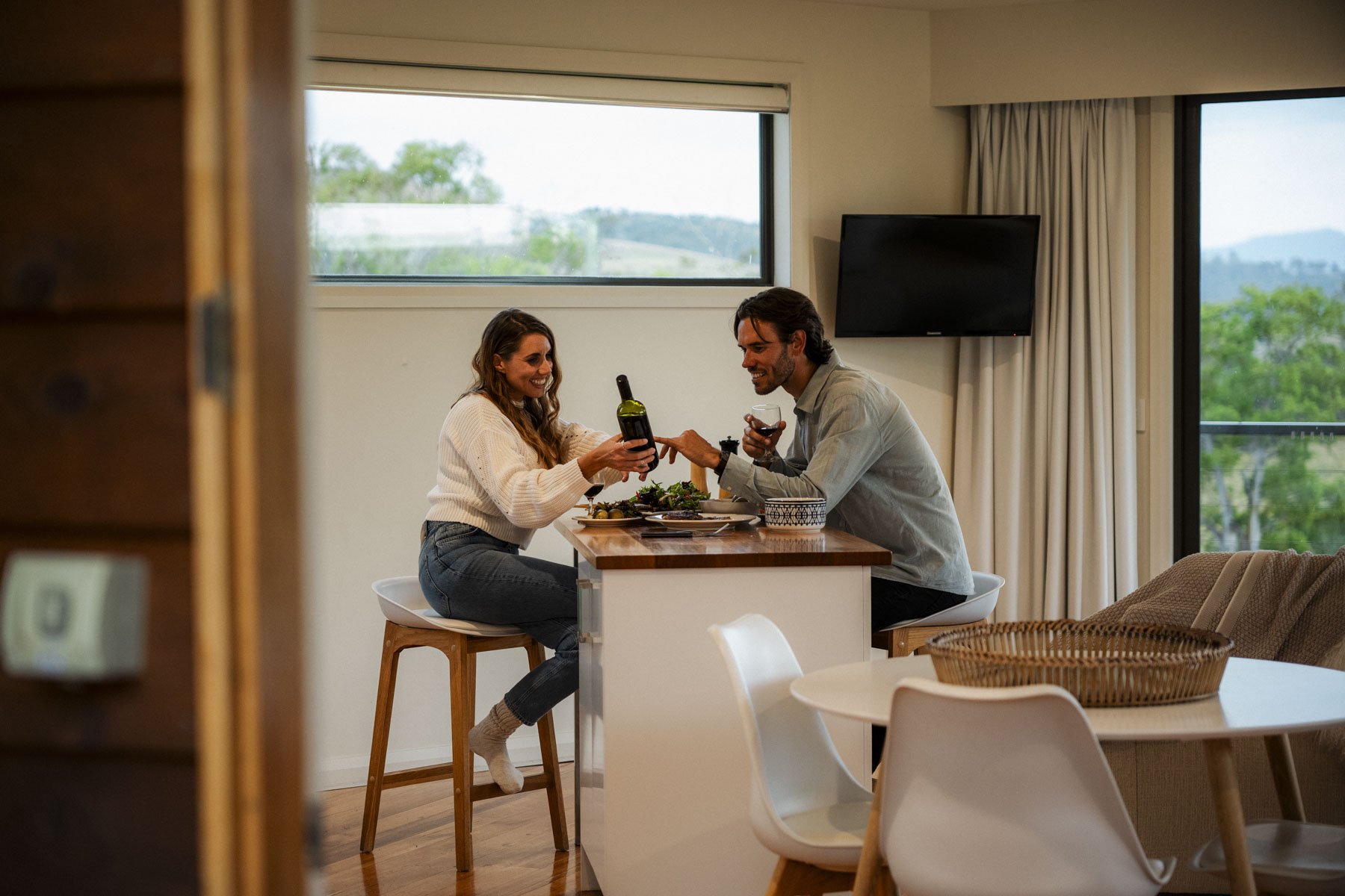 A man and a woman sitting at a kitchen island, clinking wine glasses and smiling, with a woman pouring red wine into a glass. The setting is a bright, modern kitchen with large windows showing green trees outside.