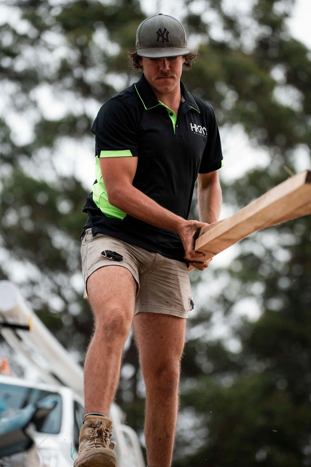 A man wearing a gray baseball cap, black and neon green shirt, and beige shorts is balancing on one foot while holding a piece of wood outdoors. He has a mustache and is standing in front of a blurred background of trees and vehicles.