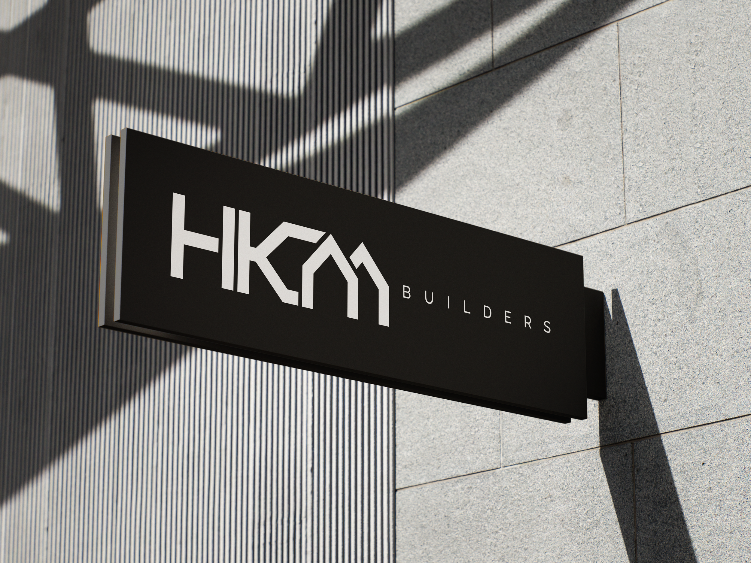 Black sign with white text reading 'HKM BUILDERS' attached to a light gray building wall with shadows cast by nearby structures.
