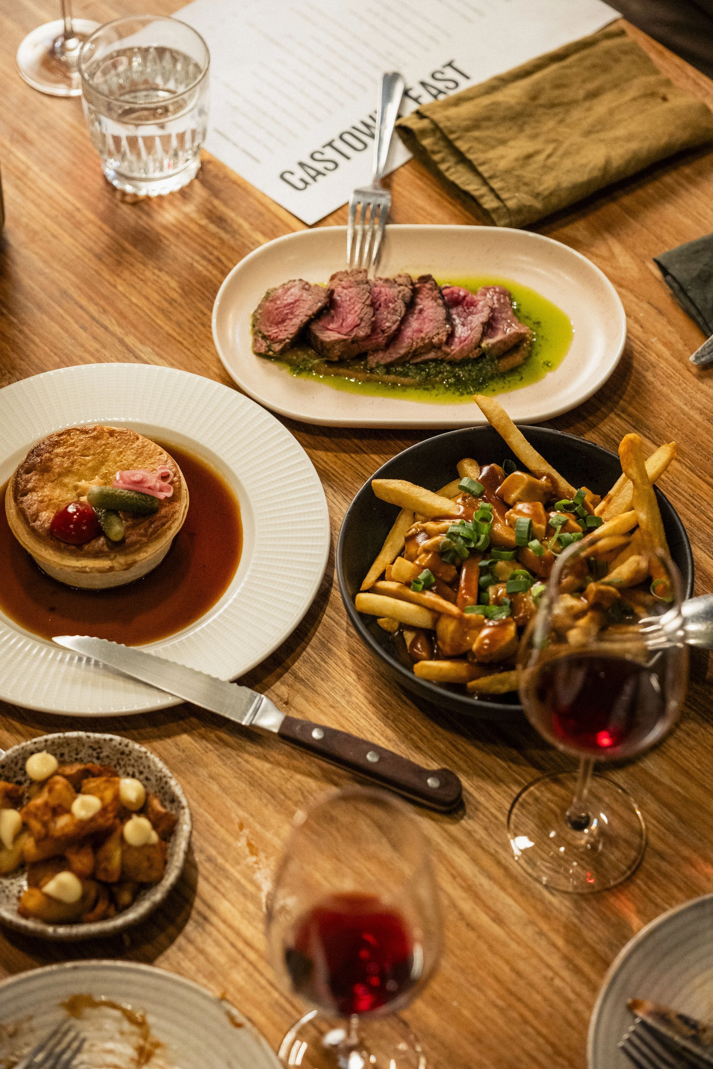 A table with a variety of dishes including sliced steak with green sauce, a meat pie in gravy, fries with gravy and chopped green onions, a small bowl of mashed sweet potatoes with white topping, and glasses of red wine.