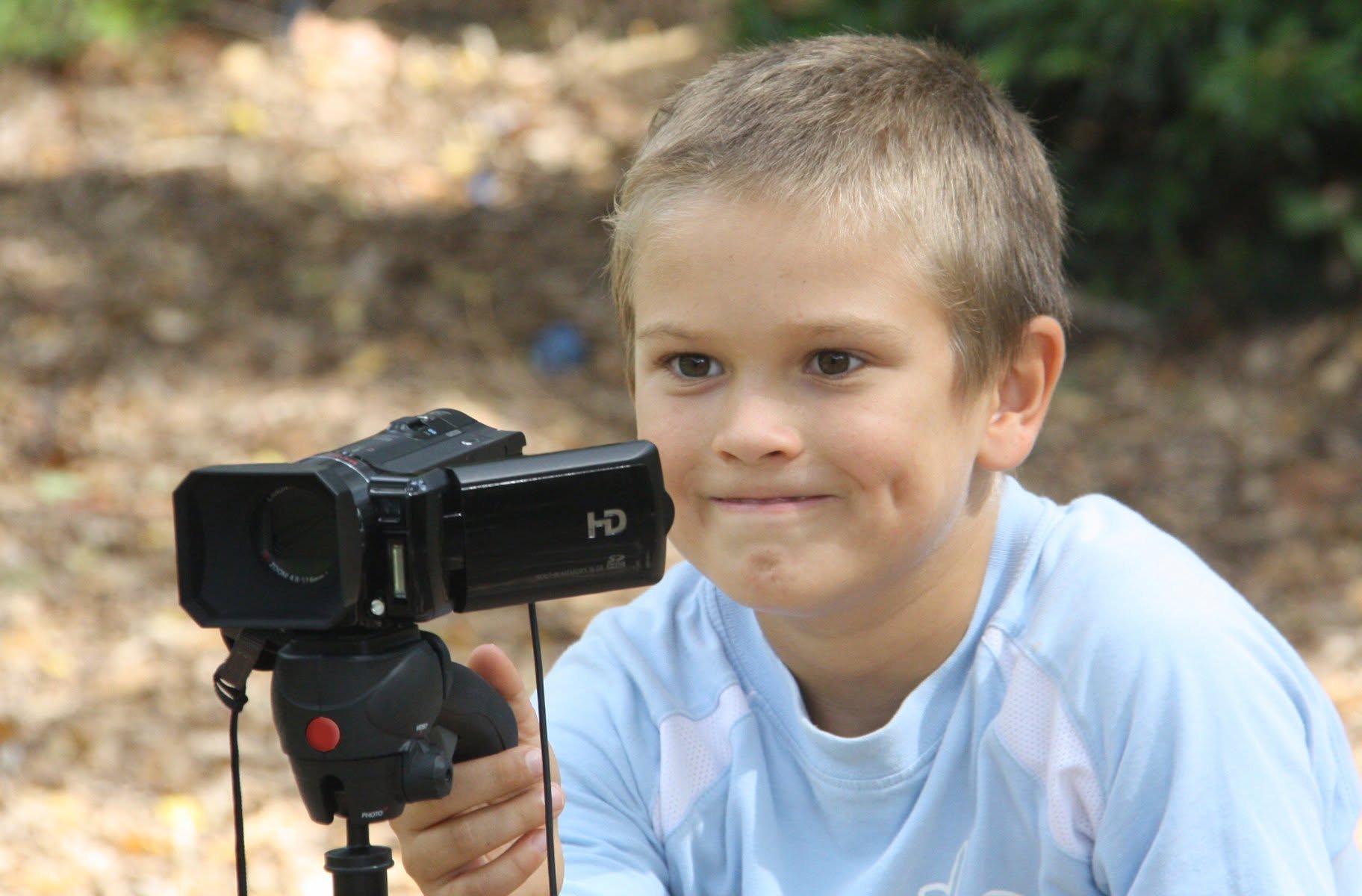 A young boy with short light brown hair looking through a camera mounted on a tripod outdoors with trees and fallen leaves in the background.