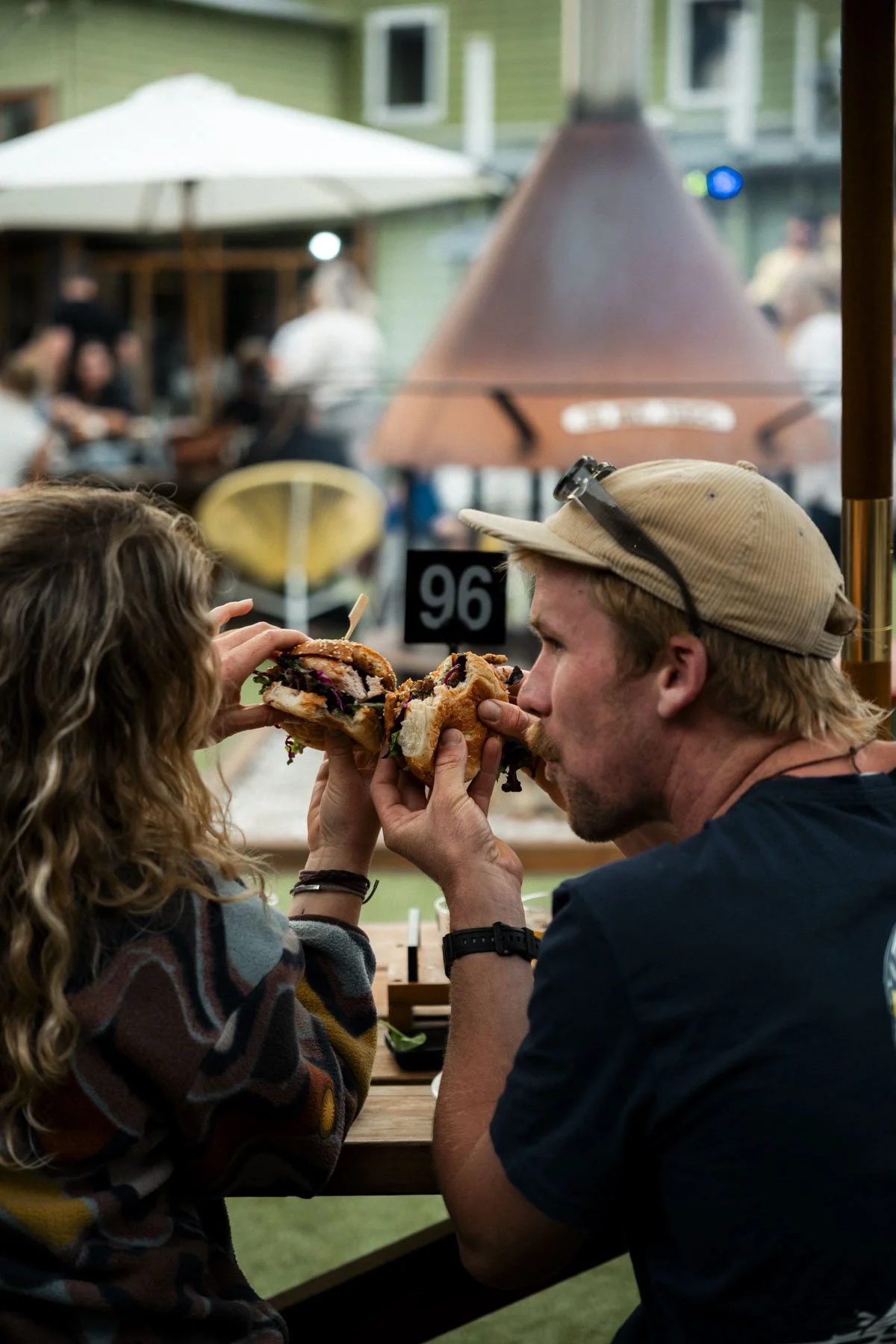 A man and woman sharing a sandwich at an outdoor restaurant or cafe. There are people and umbrellas in the background.