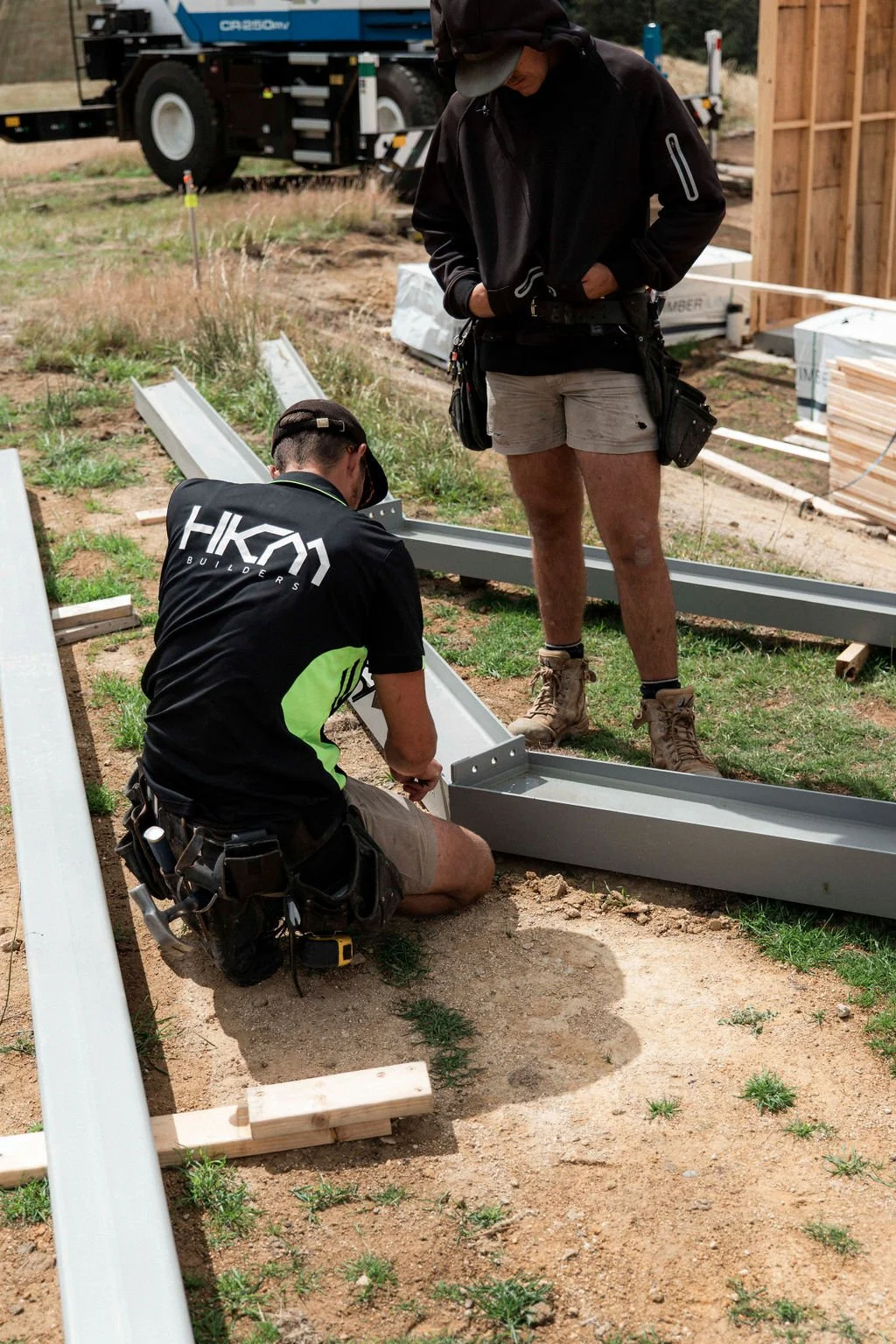 Two construction workers assembling a metal frame on the ground at a construction site. One worker is kneeling, wearing a black and green shirt, and the other is standing, wearing shorts and a black jacket. Construction materials and equipment are visible around them.
