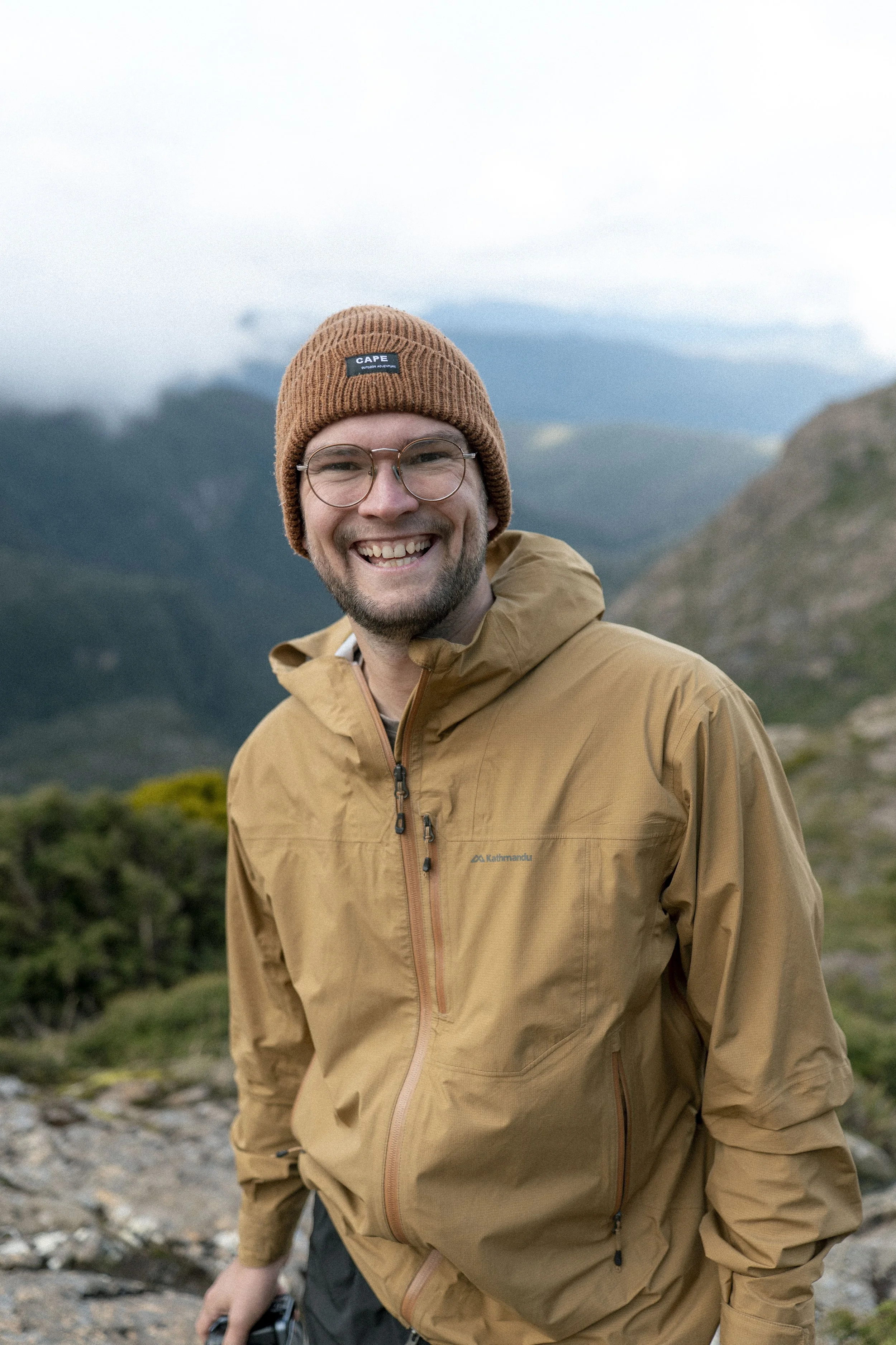 A smiling man wearing glasses, a brown beanie, and a tan outdoor jacket standing in a mountainous landscape with cloudy sky and green hills in the background.