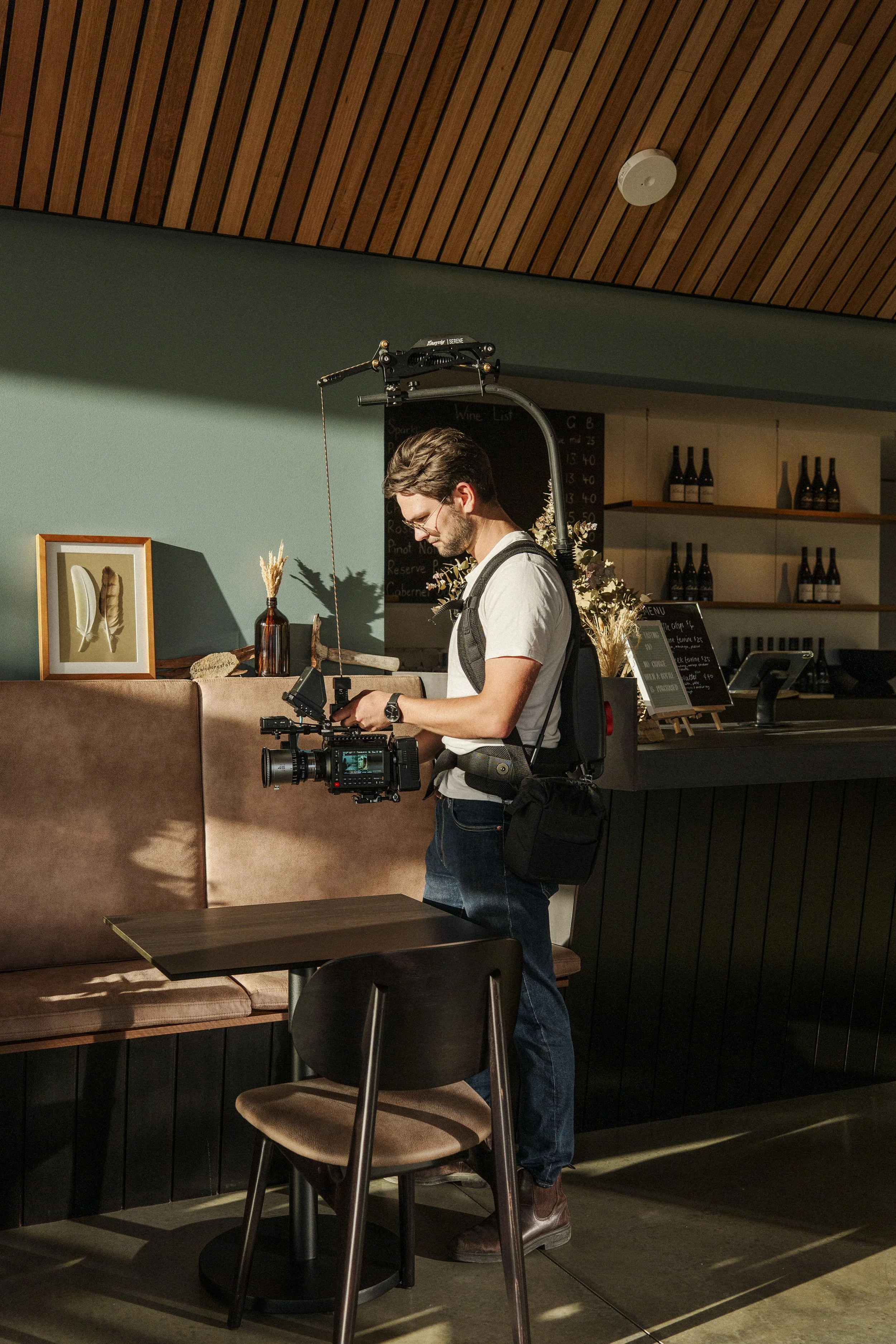 A man filming with professional video equipment inside a cafe or restaurant, with shelves of wine bottles in the background.