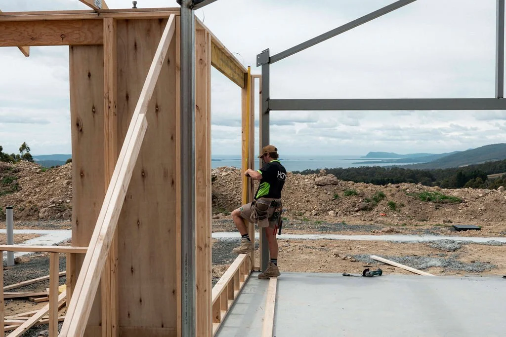 A construction worker working on a wooden building frame at a construction site with a scenic landscape in the background.