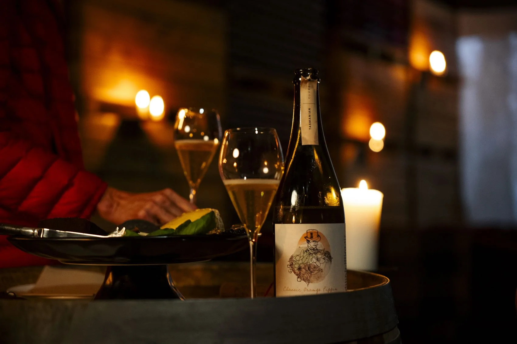 A bottle of sparkling wine or champagne, two filled glasses, a plate with food, and lit candles on a table in a dimly lit setting.