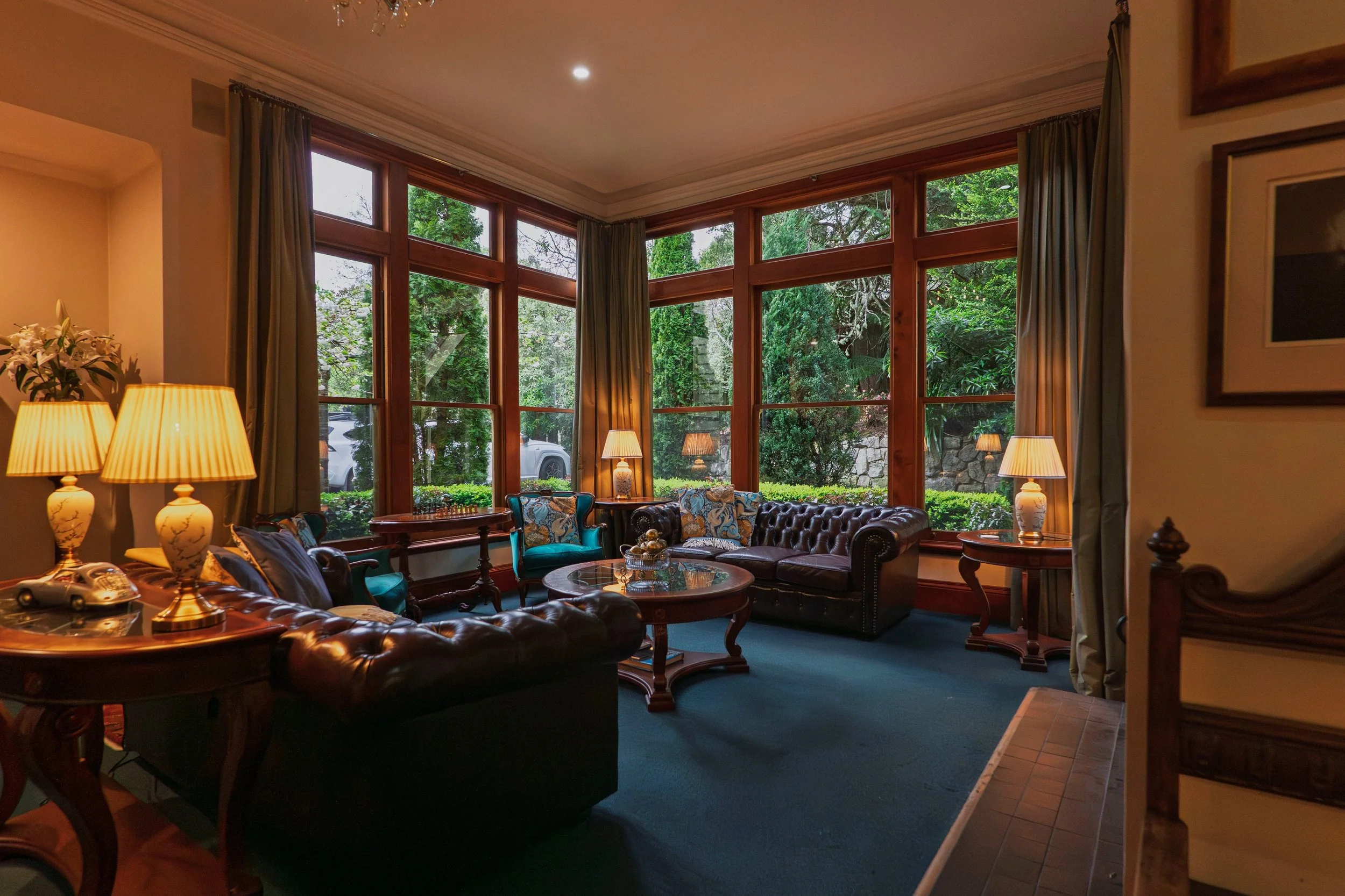 A cozy living room with large wooden-framed windows overlooking greenery. Contains leather sofas, patterned armchairs, wooden side tables with lamps, and decorative items.