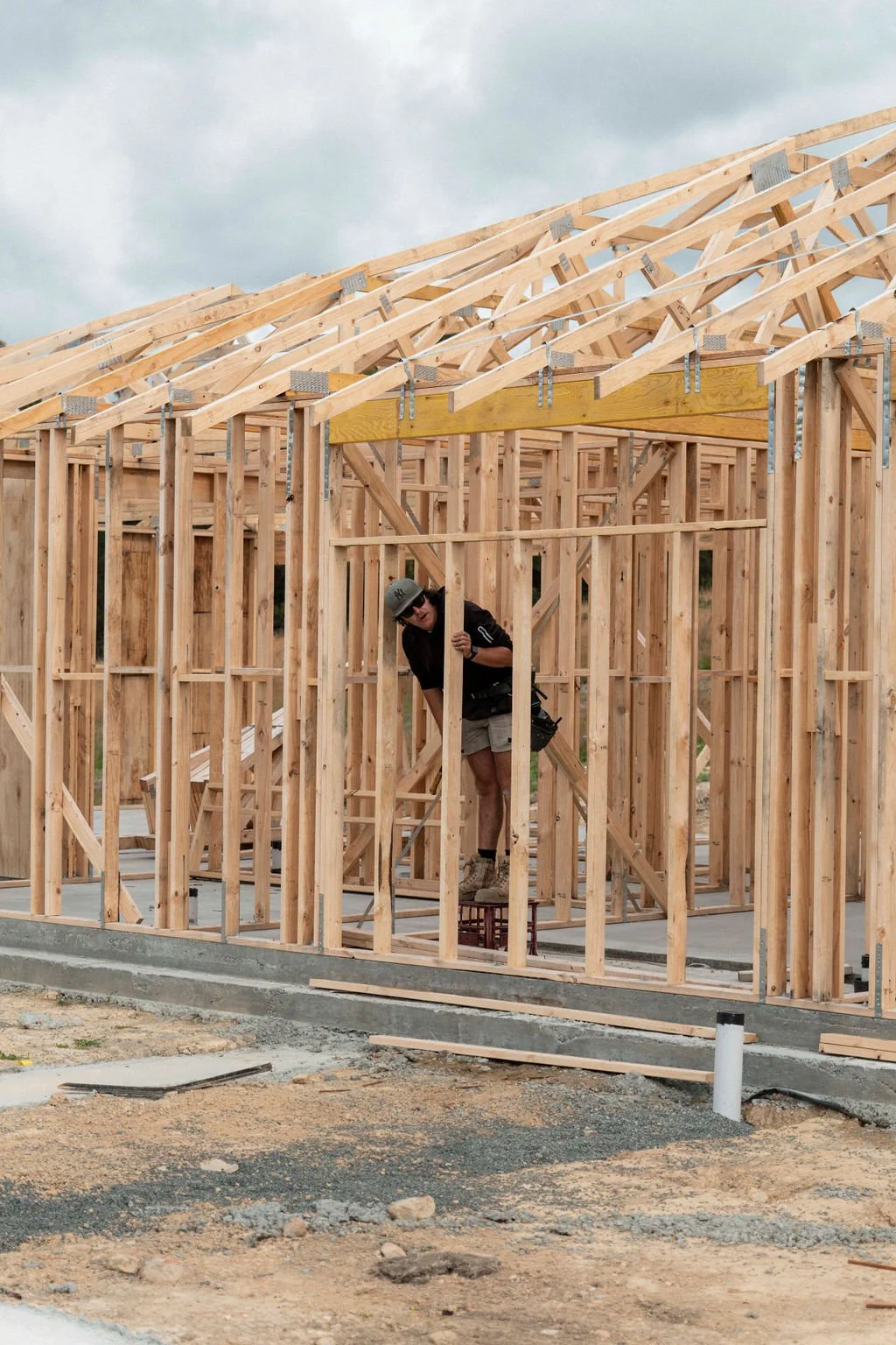 A construction worker wearing a helmet and shorts is inside a wooden framework of a house under construction, working on the structure.