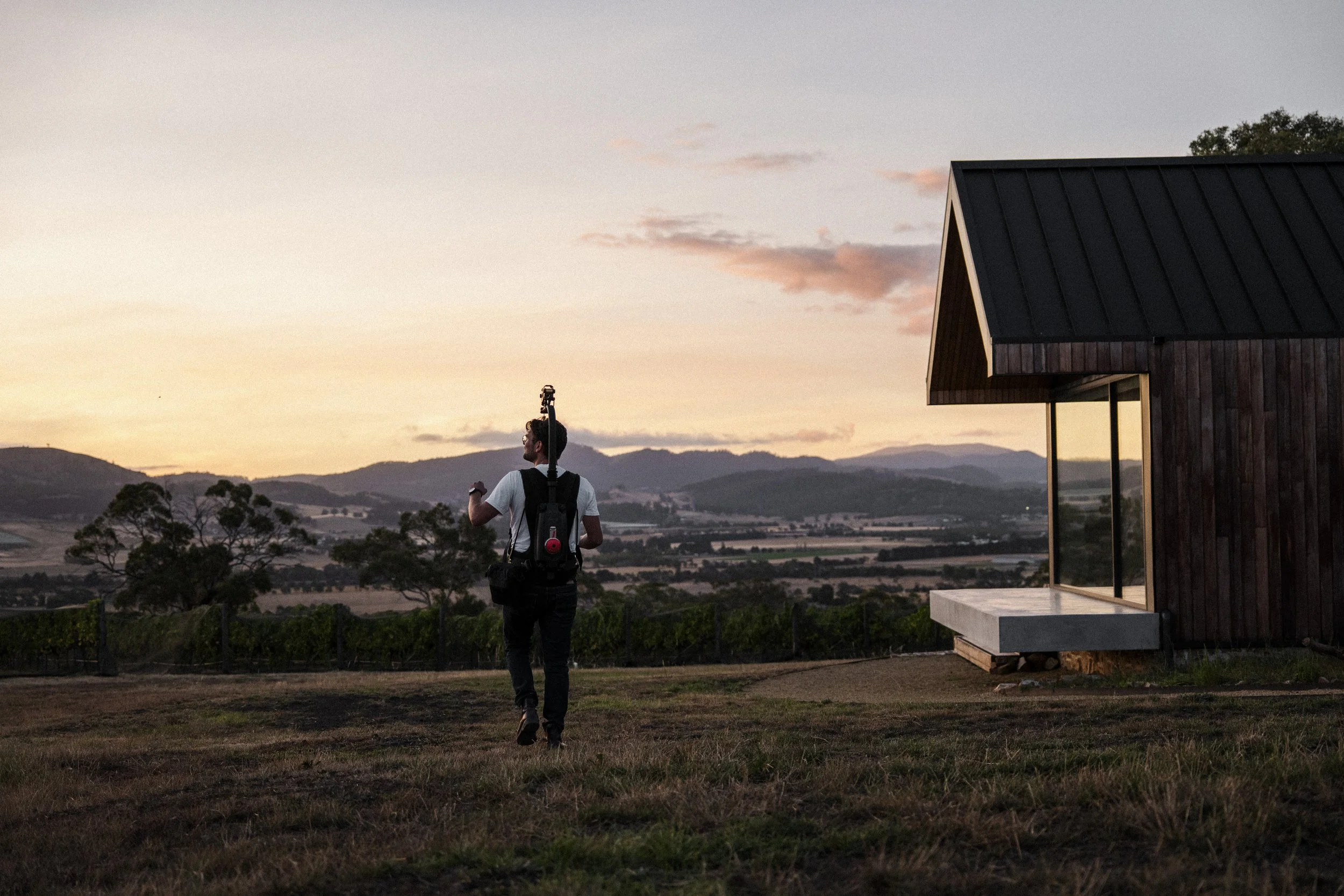 A person with a backpack and tripod walking near a modern wooden house during sunset in a rural landscape with hills and trees.