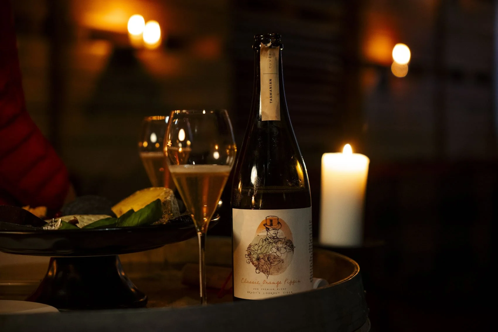 A bottle of cider labeled "Classic Orange Pippin" with two glasses of champagne in a dimly lit setting with candles.