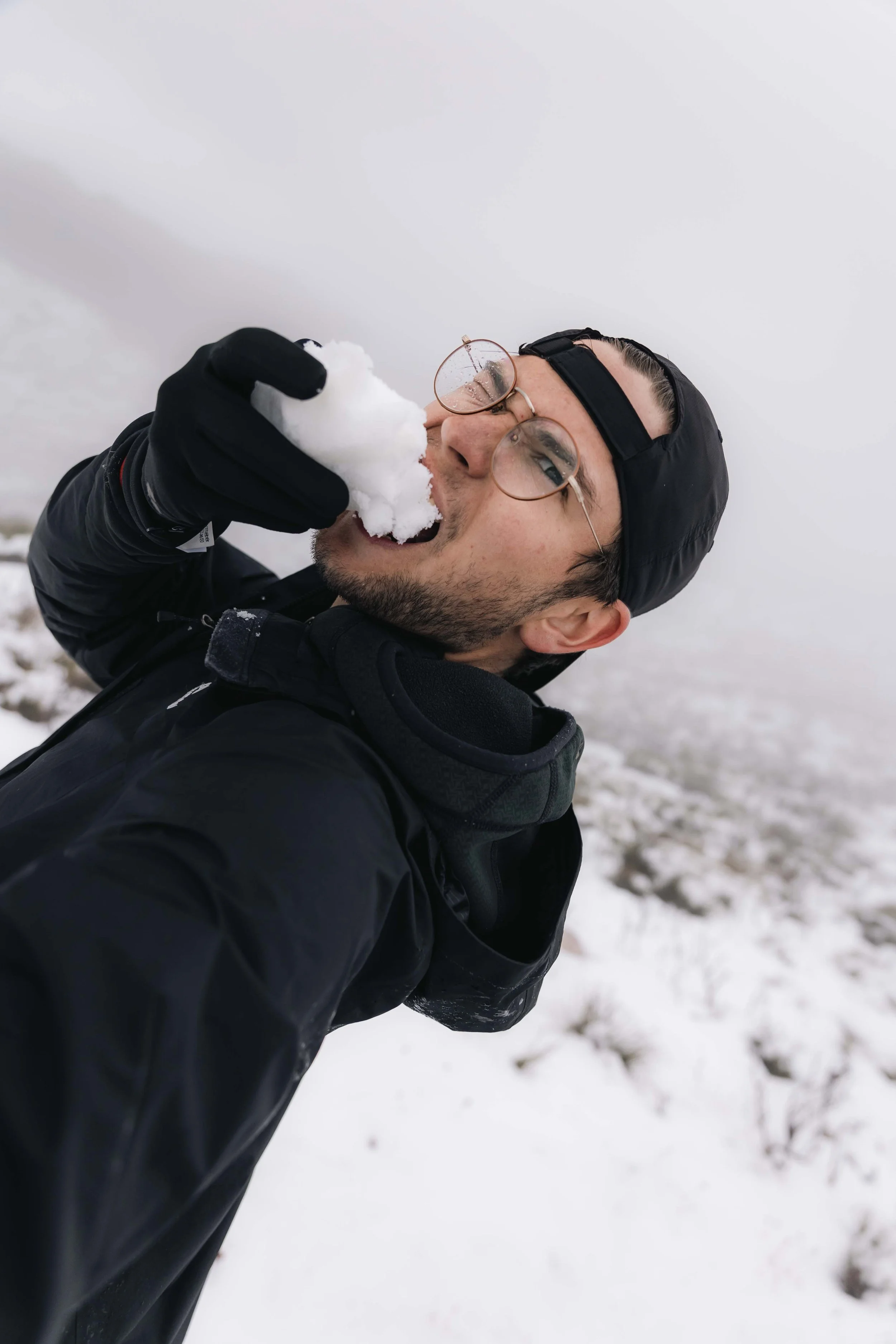 A man wearing glasses, a black jacket, and a black cap is outdoors in snowy weather, holding and biting a piece of snow.