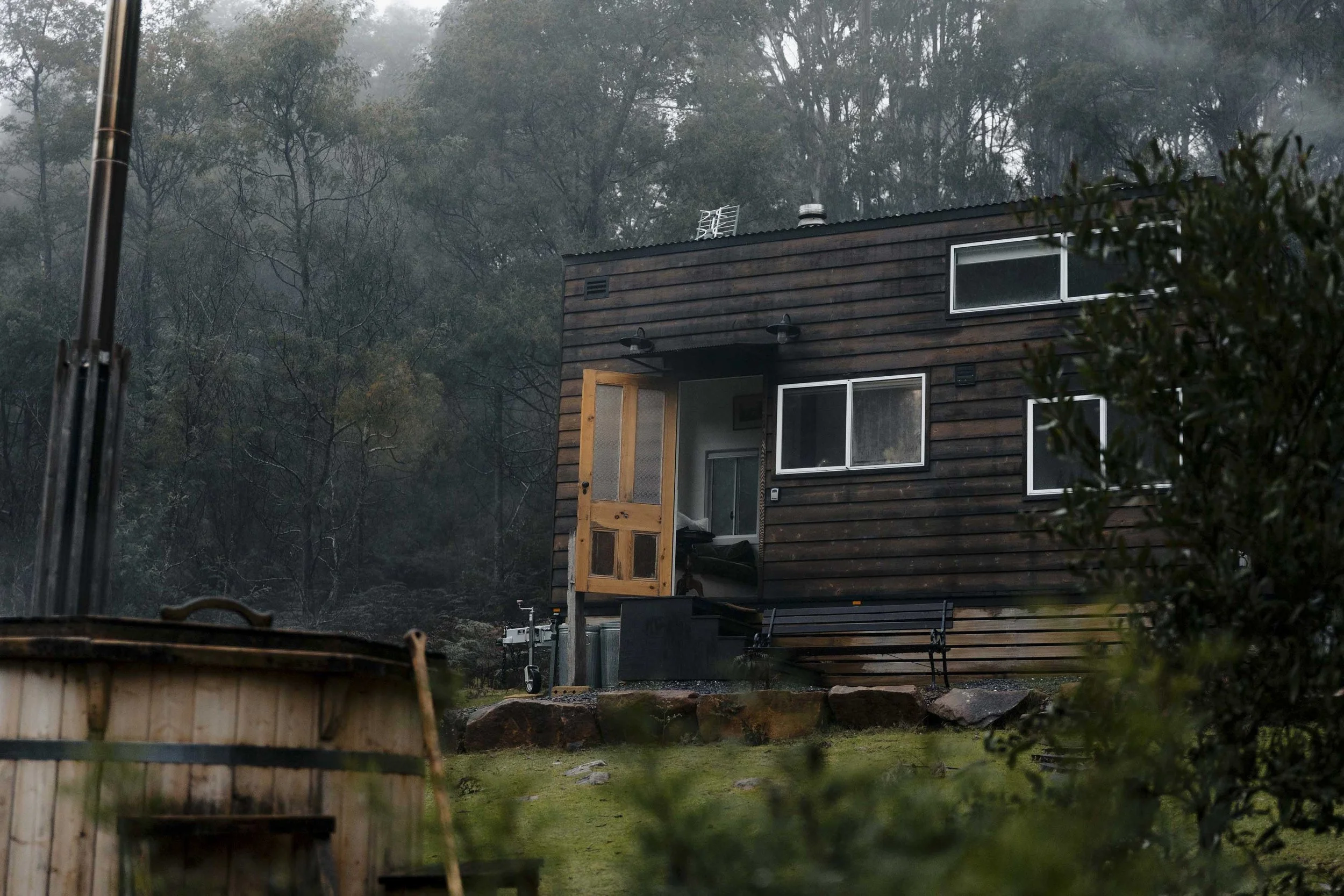 A dark wooden house with multiple windows and an open front door, surrounded by greenery and trees, with a bench and outdoor equipment nearby.