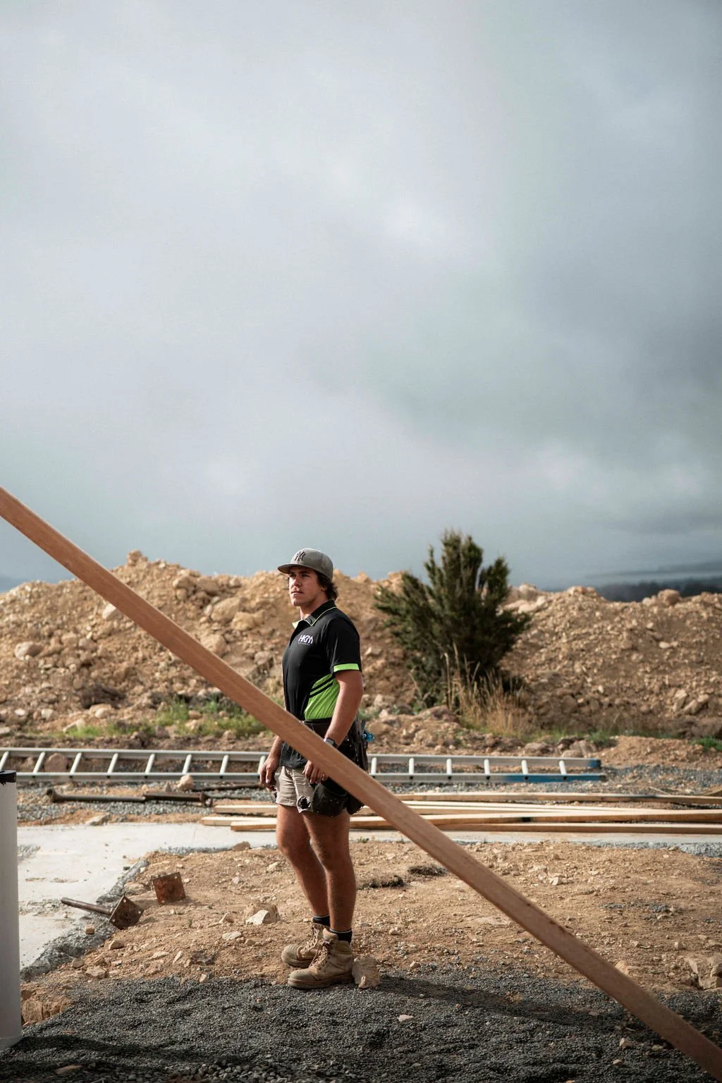 A young man standing on a construction site outdoors, wearing work boots, shorts, and a black polo shirt, with overcast skies and dirt mounds in the background.