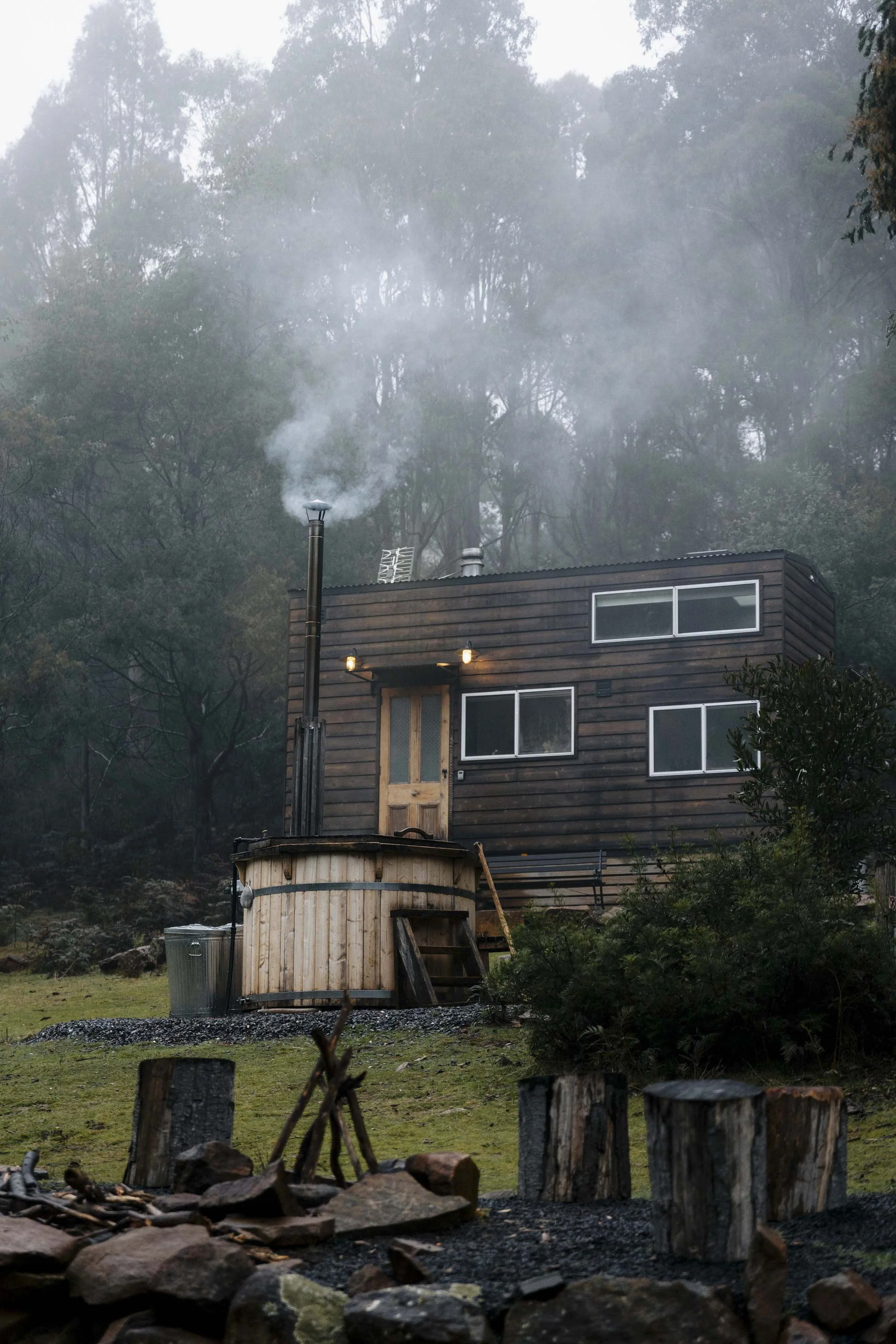 A small modern house with wood siding and large windows, surrounded by trees and fog, with a hot tub in front emitting steam.