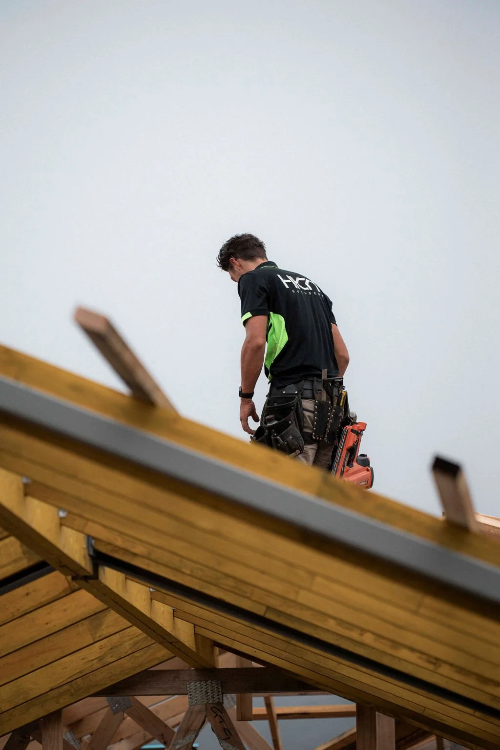 A construction worker wearing a black and green shirt with tool belt working on a wooden roof structure.
