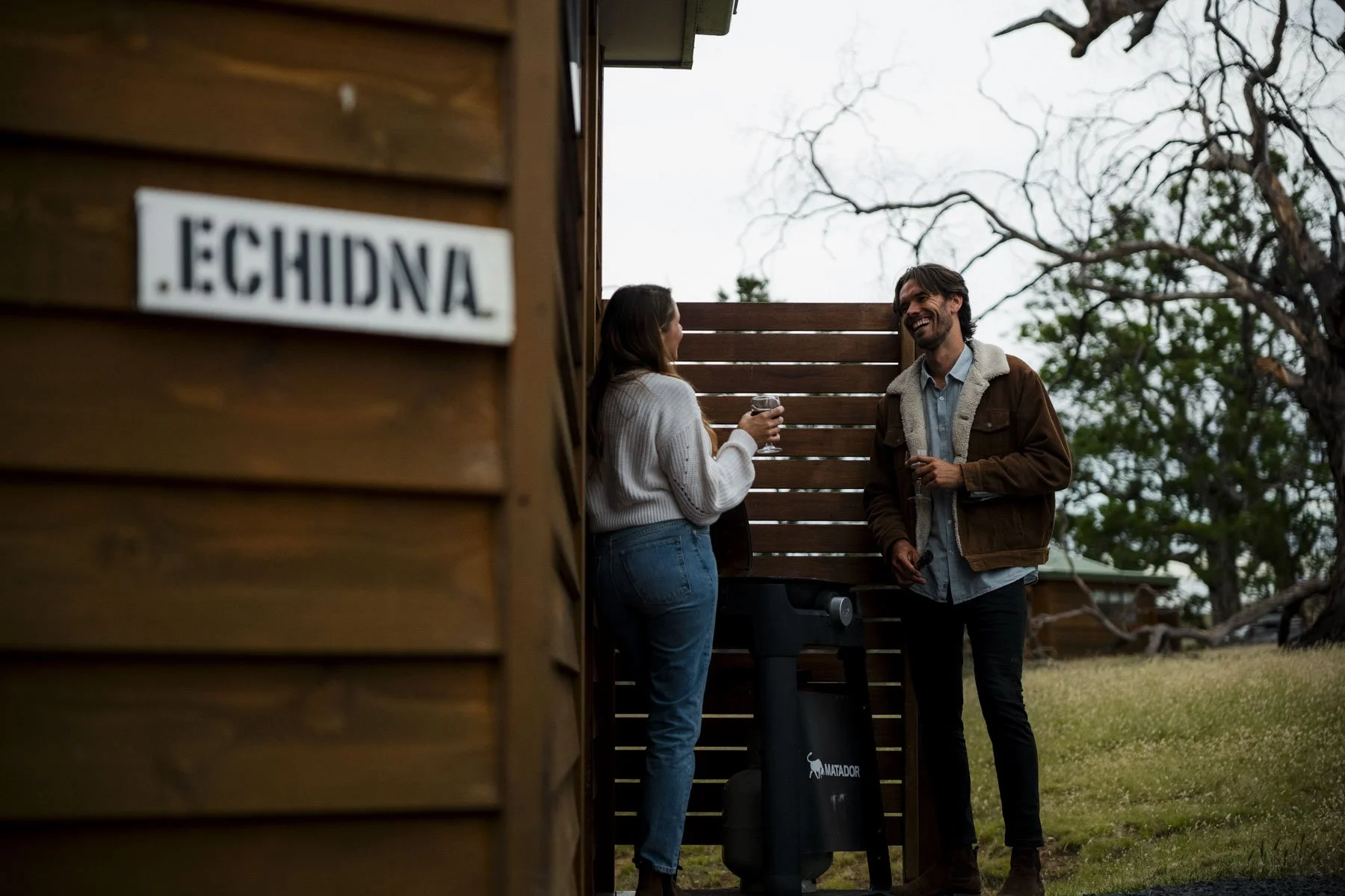 Two people enjoying drinks and talking outdoors near a wooden structure with a sign that reads 'Echidna'; one person is a woman in a white sweater and blue jeans, the other is a man in a brown jacket and black pants, smiling and laughing.