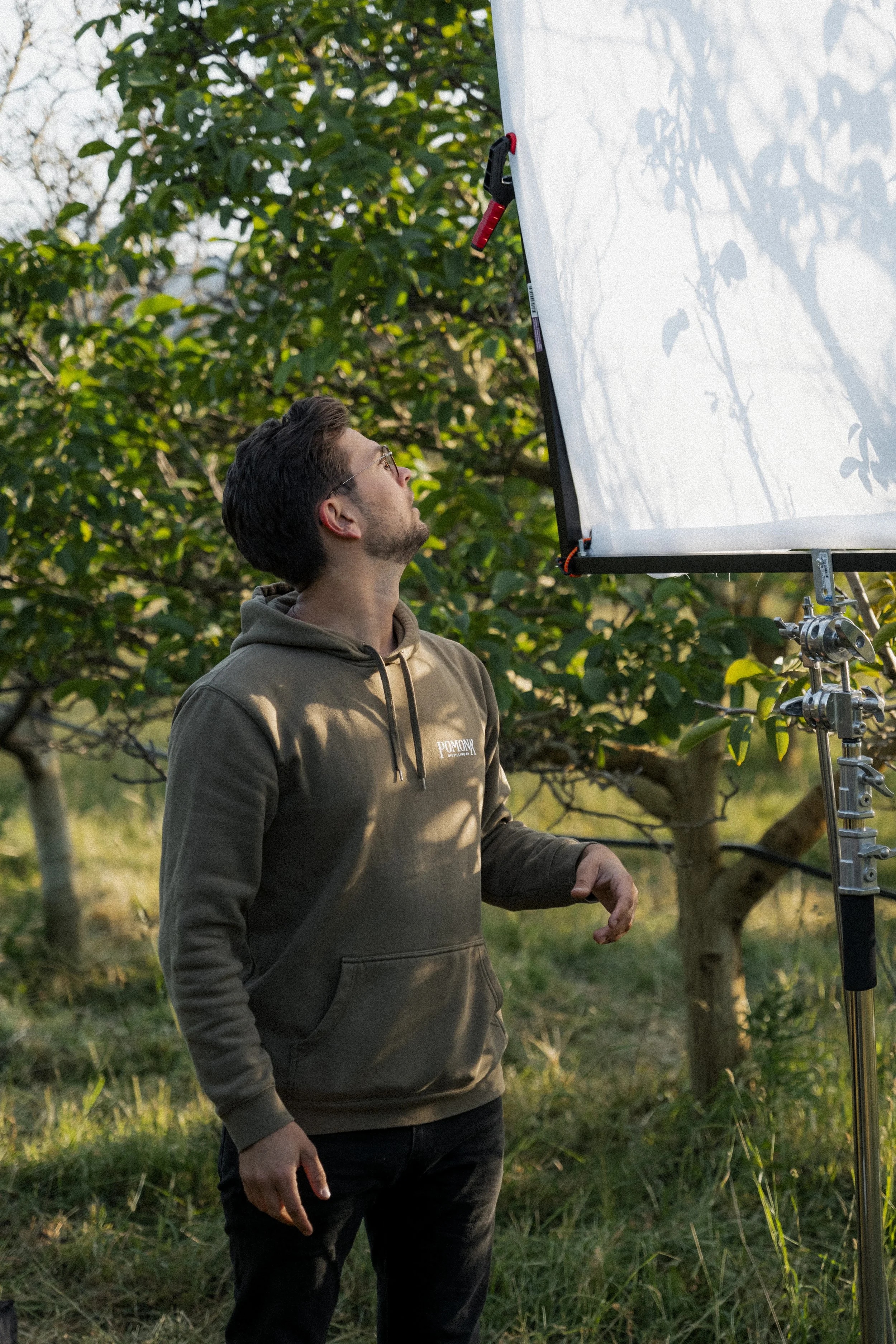 Man in hoodie standing outdoors, looking up at a large light reflector with trees in the background.