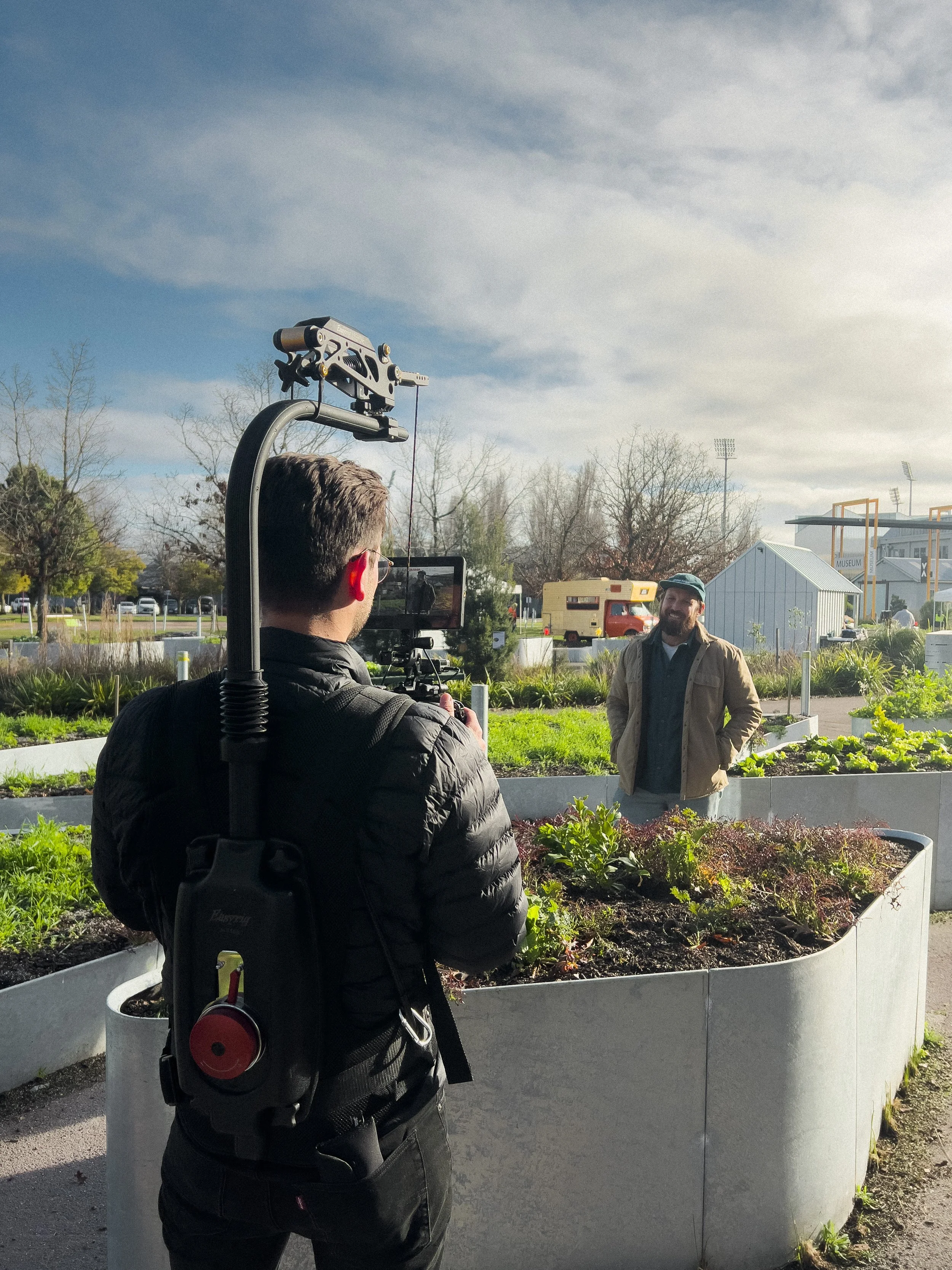A man being filmed by a camera operator in an outdoor garden or park area under a partly cloudy sky.